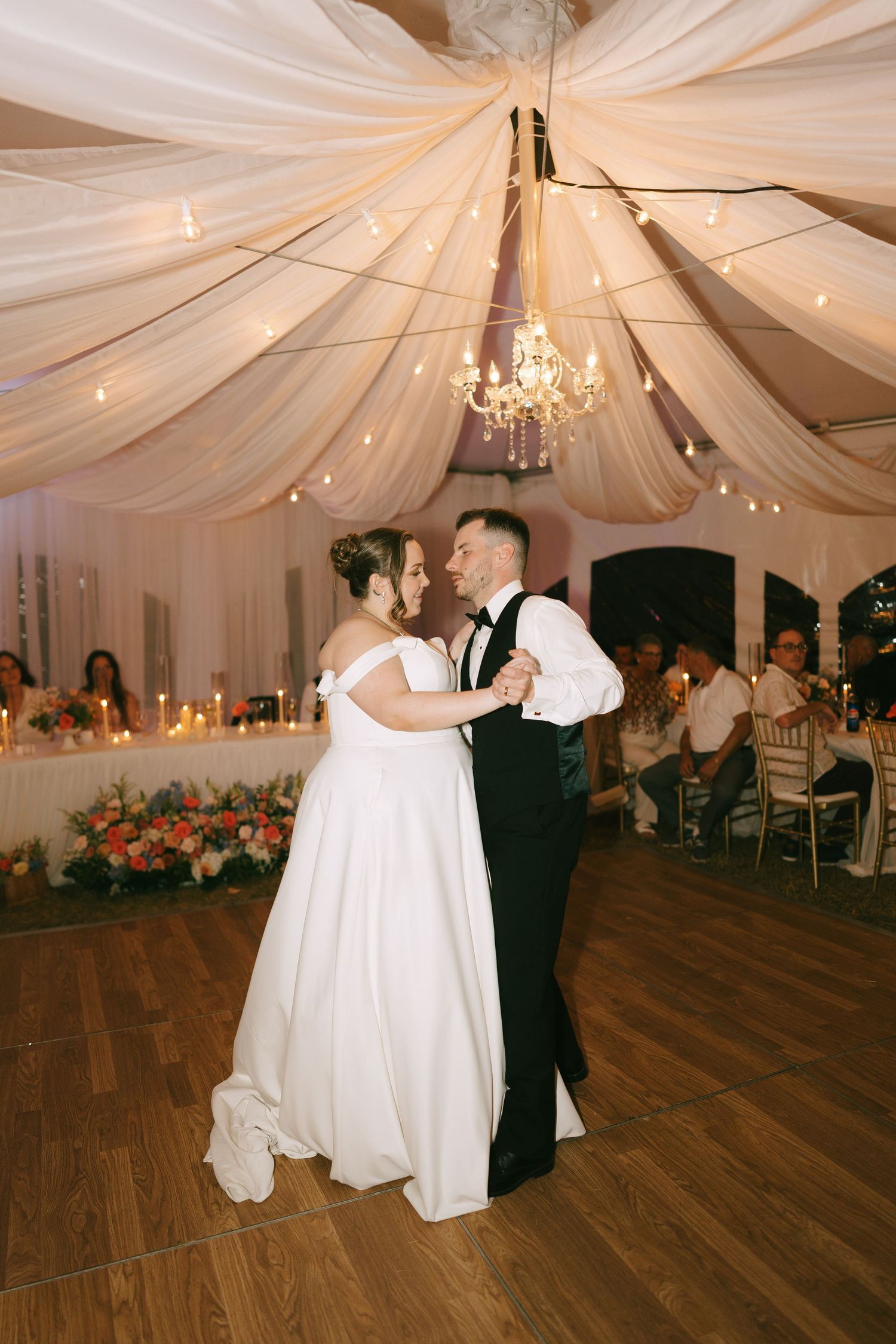A couple performs a romantic dance move in an elegant white-draped reception tent with chandeliers.