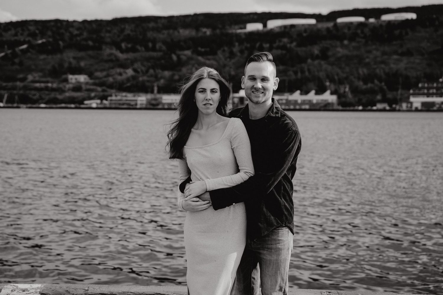 A seaside photoshoot featuring a couple on a concrete walkway with water and hills behind them.
