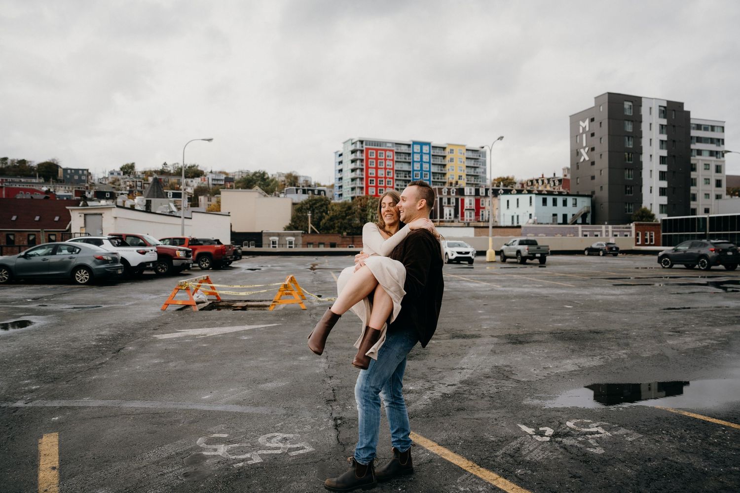 A couple dances playfully on a city rooftop parking lot with urban buildings in the background.
