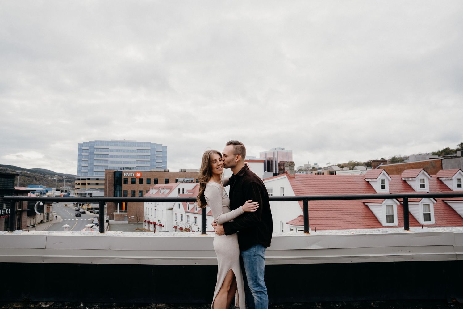A couple shares a romantic moment on a rooftop with modern buildings and red roofs in the background.