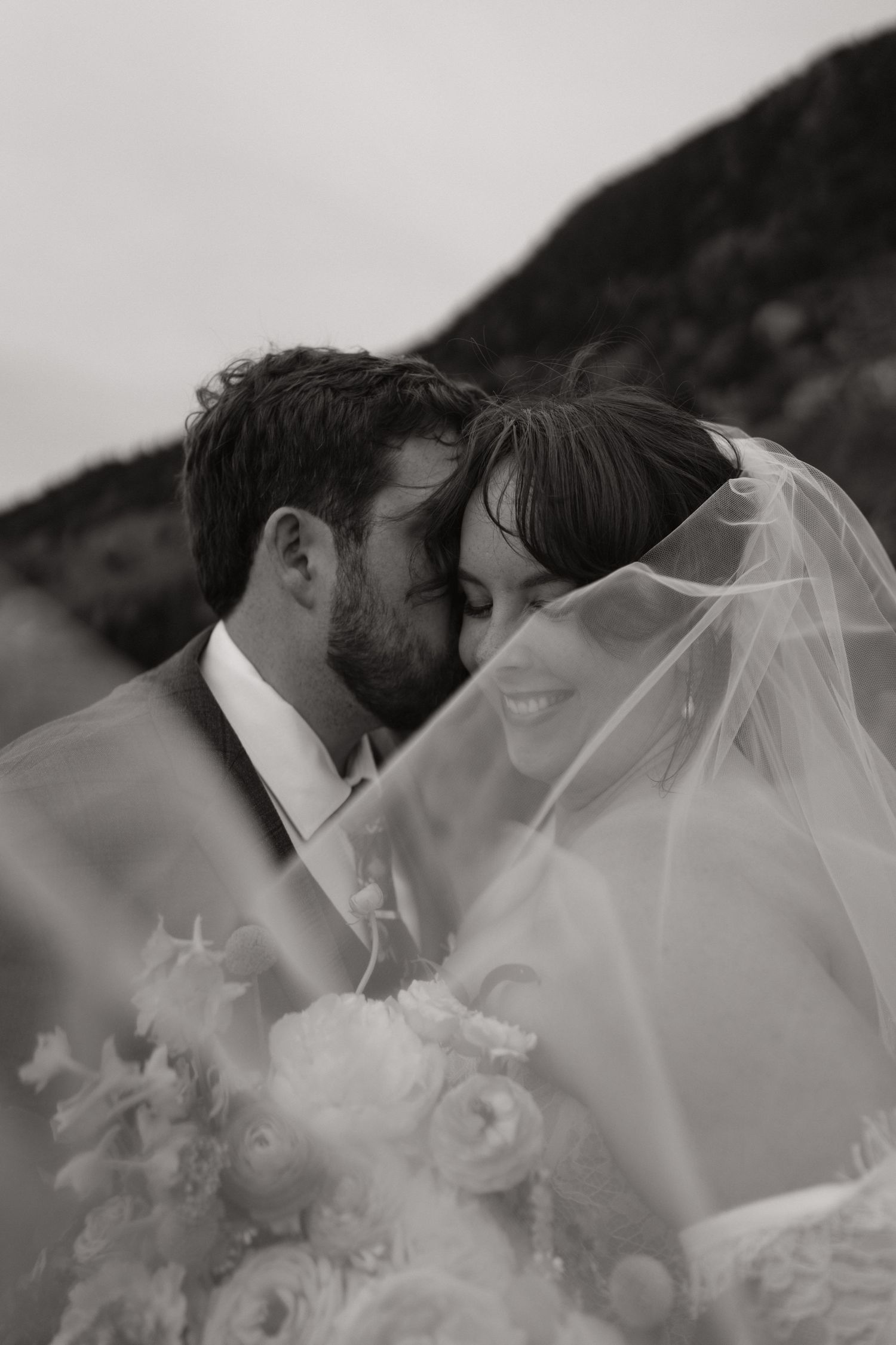 Black and white wedding photo of bride and groom kissing under veil with bridal bouquet outdoors.