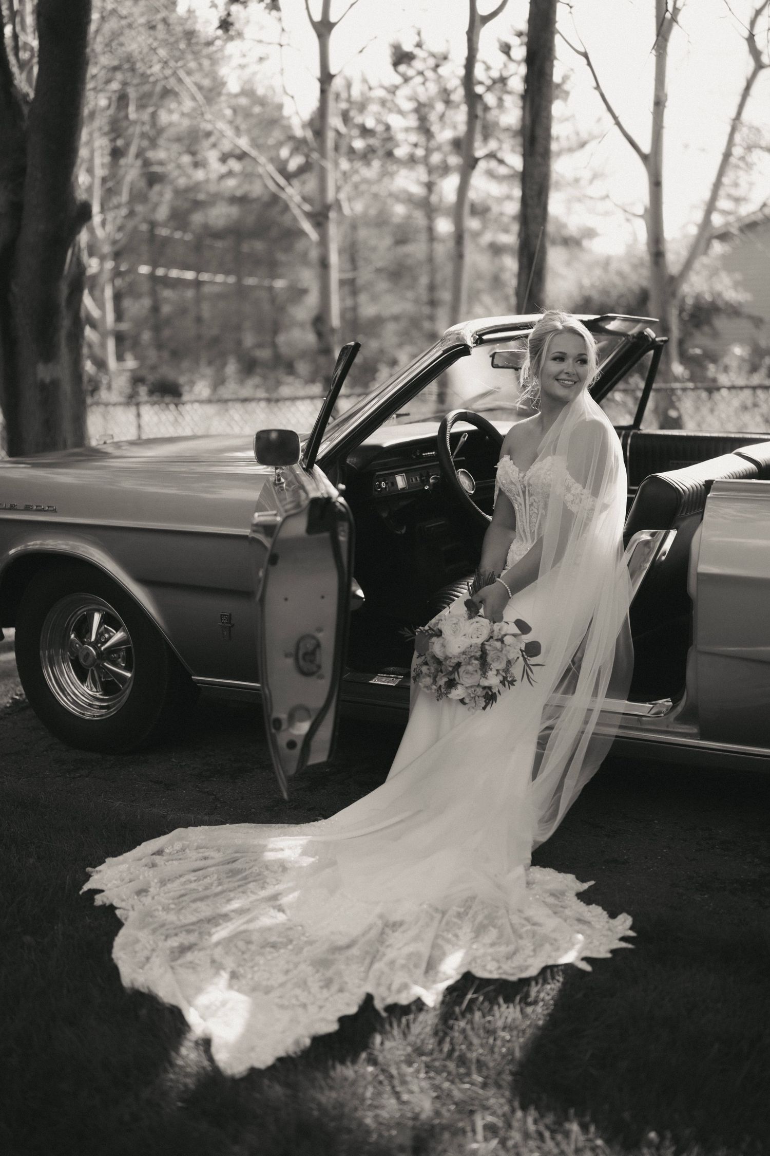 Elegant bride in a lace wedding gown and veil poses beside a vintage convertible car, holding a bouquet, in black and white.