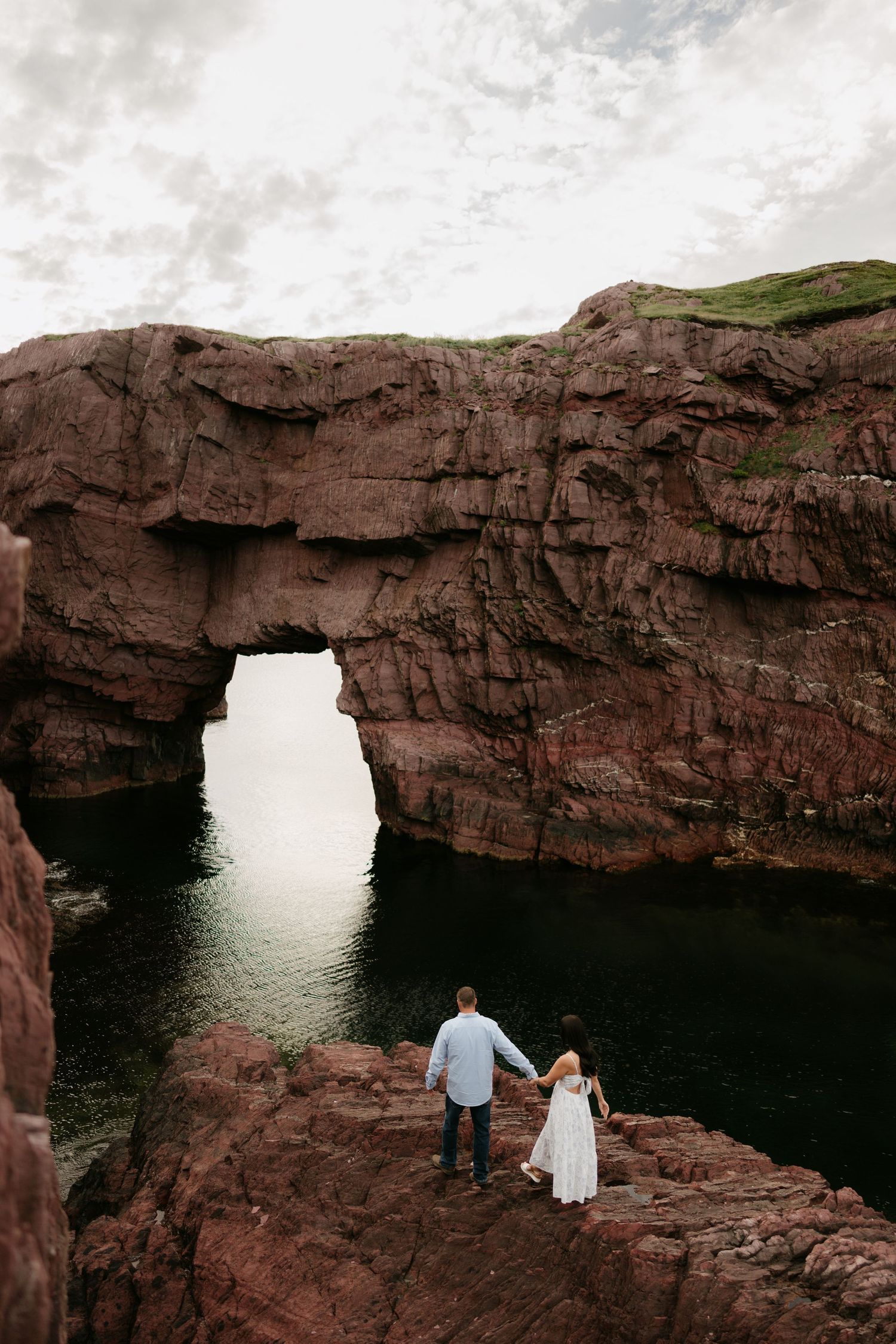 Couple holding hands on red rock cliffs beside a natural stone arch over calm green water.