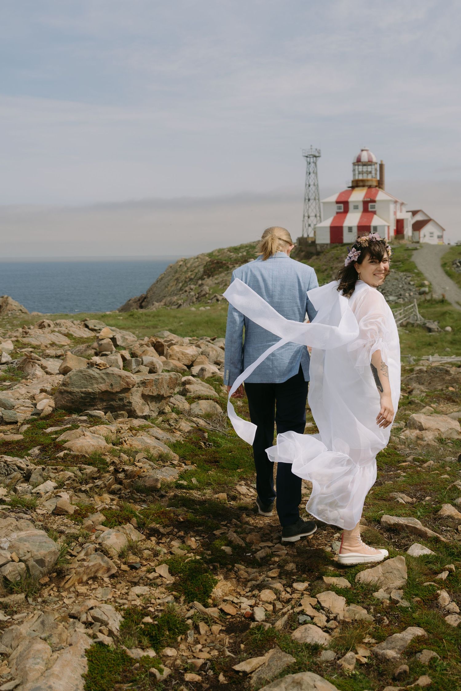 A white dress flows in the wind against a rocky coastal landscape with a lighthouse in the background.