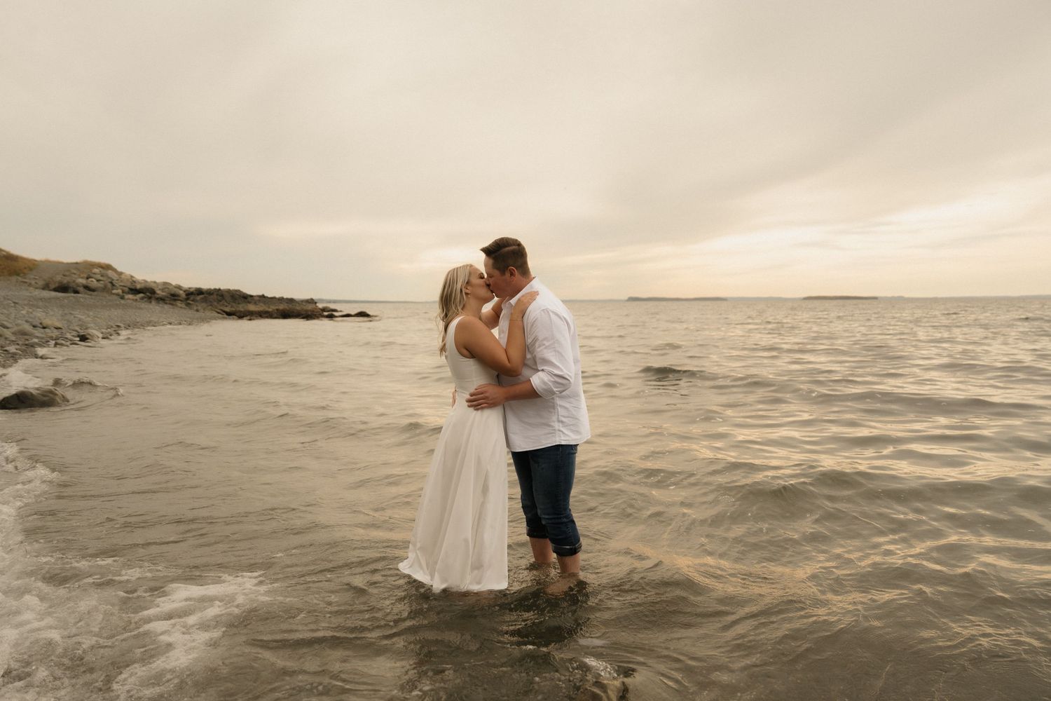 A romantic couple embraces at sunset on a beach while waves gently crash around them in the ocean.