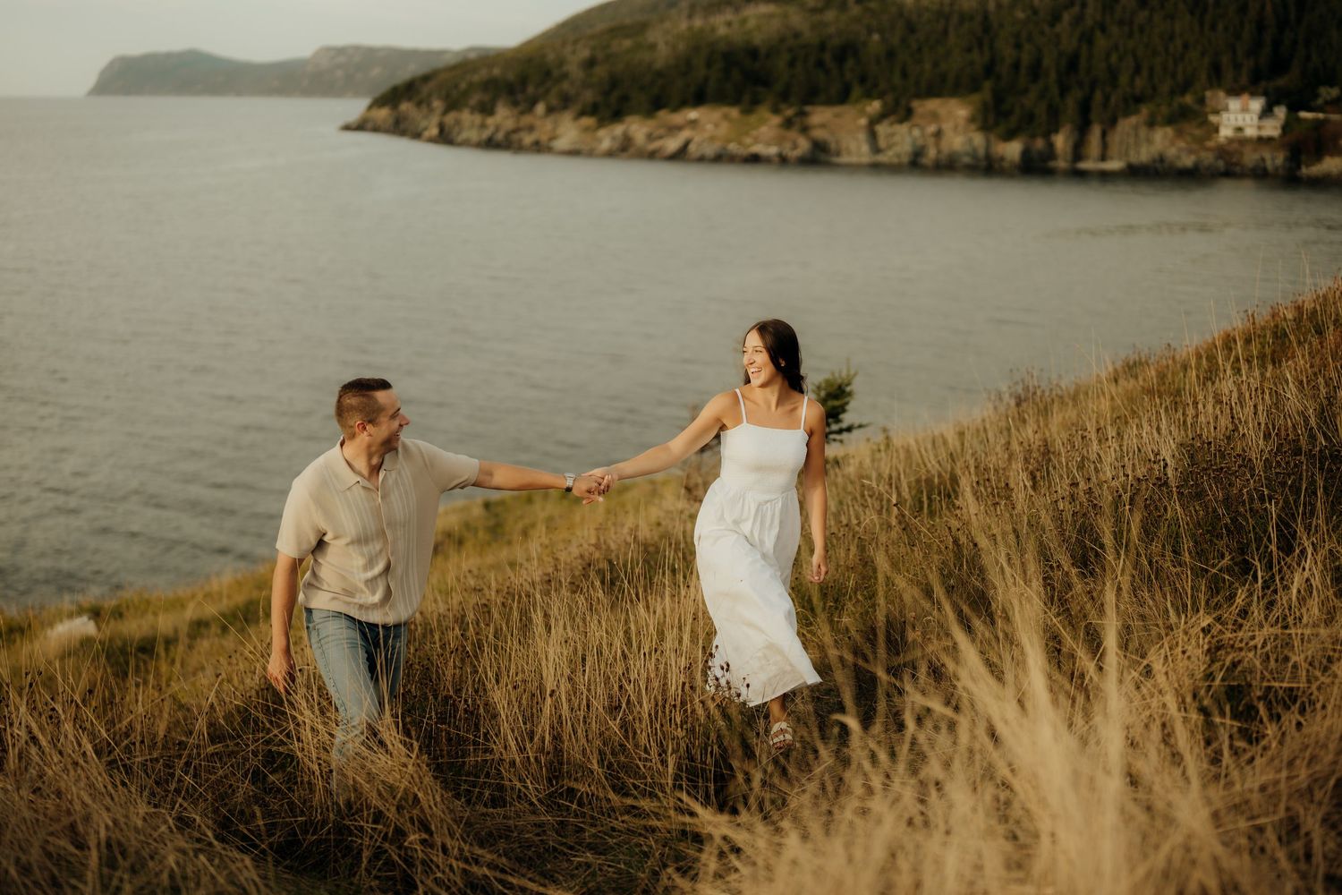 Couple walking through golden grass along coastal cliffs at sunset with ocean views in background.