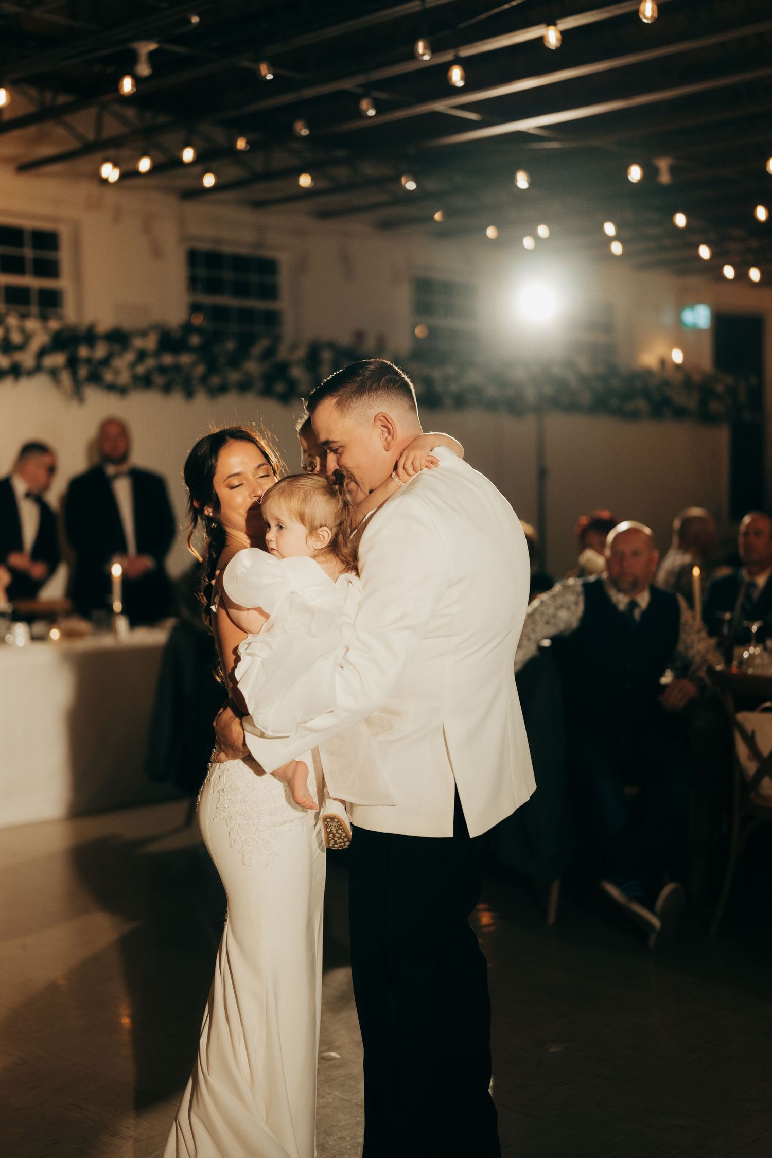 A couple shares an intimate dance moment at their wedding reception under warm string lights in an indoor venue.