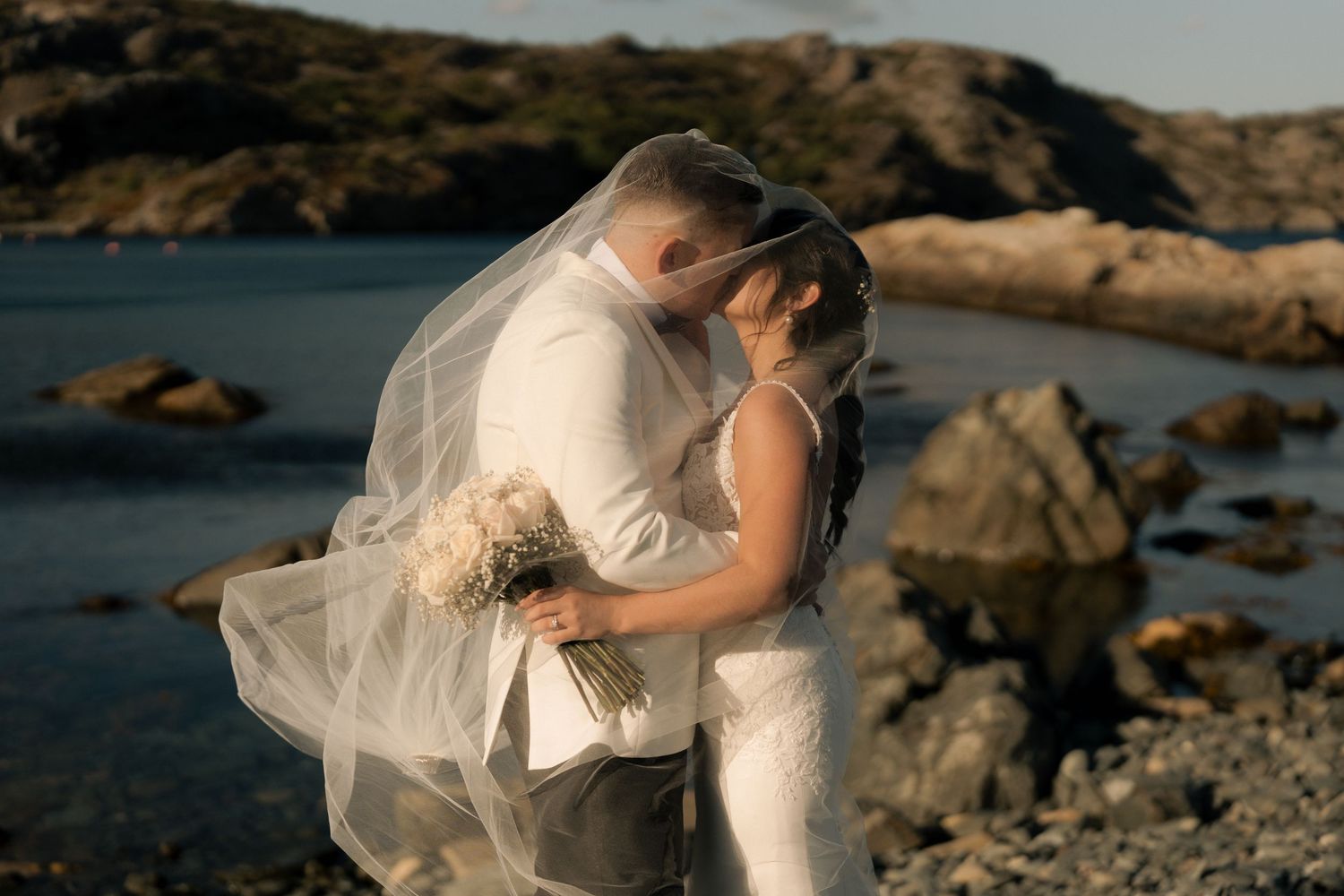 A romantic wedding photo session on a rocky beach coastline with flowing white dress and dramatic ocean backdrop.