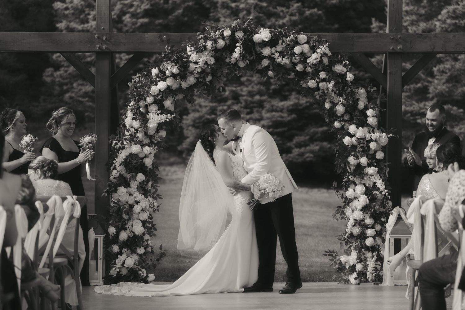 A black and white panoramic wedding ceremony scene in an outdoor garden setting with floral decorations.