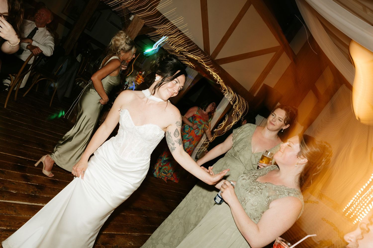 Bride in white gown dancing with guests in elegant gowns at a rustic wedding reception venue.