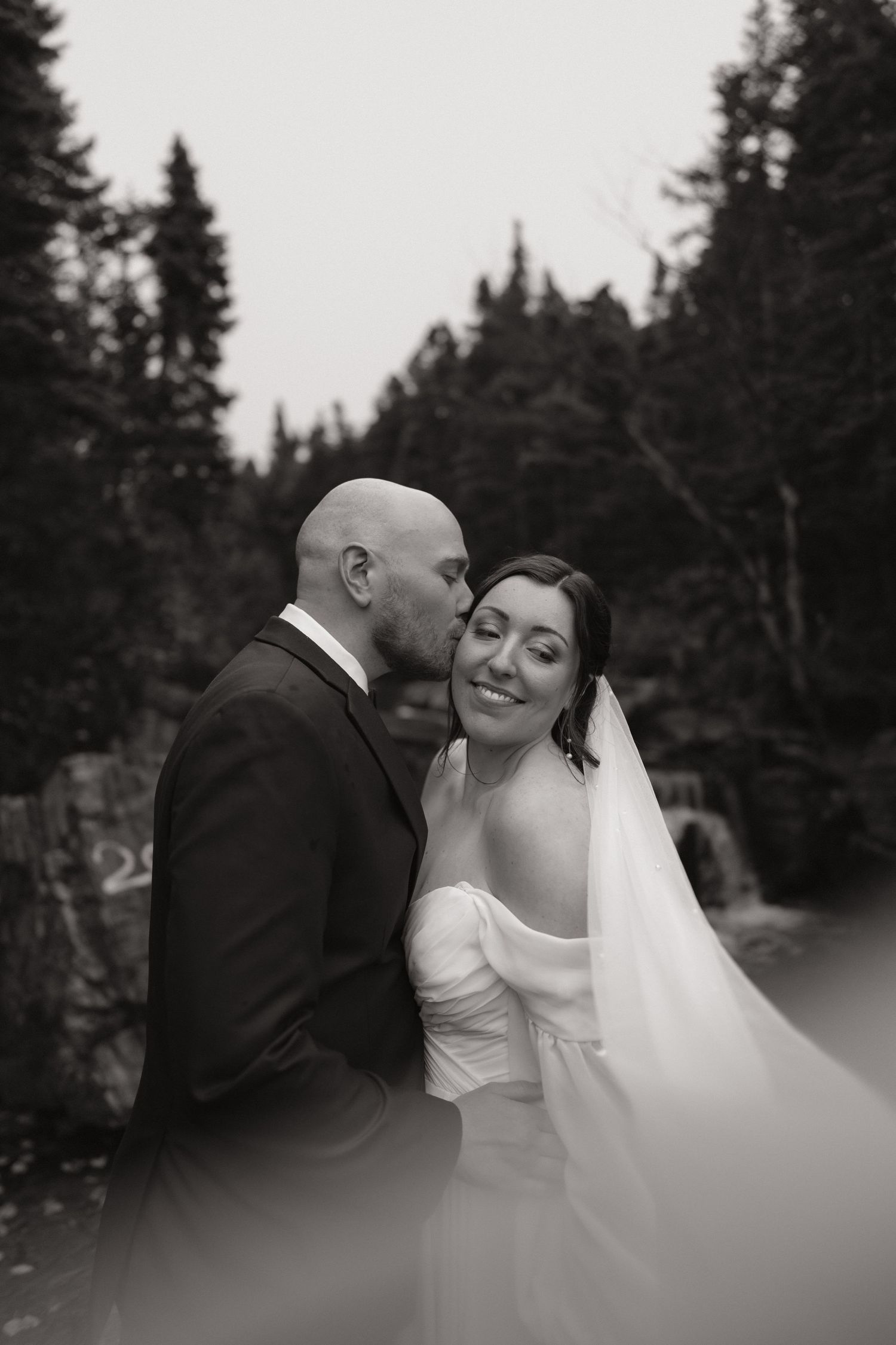 A tender black and white wedding moment captured in a forest setting with evergreen trees in the background.