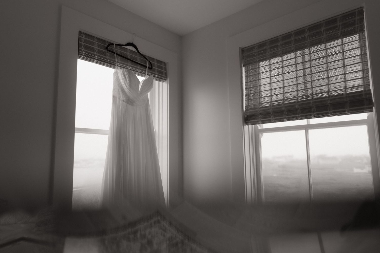 A white wedding dress hangs by a window in a minimalist room, captured in black and white photography.