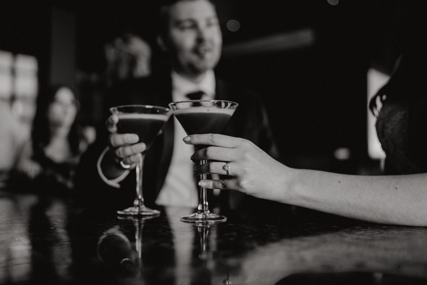 Two hands toasting with martini glasses at a dimly lit bar.