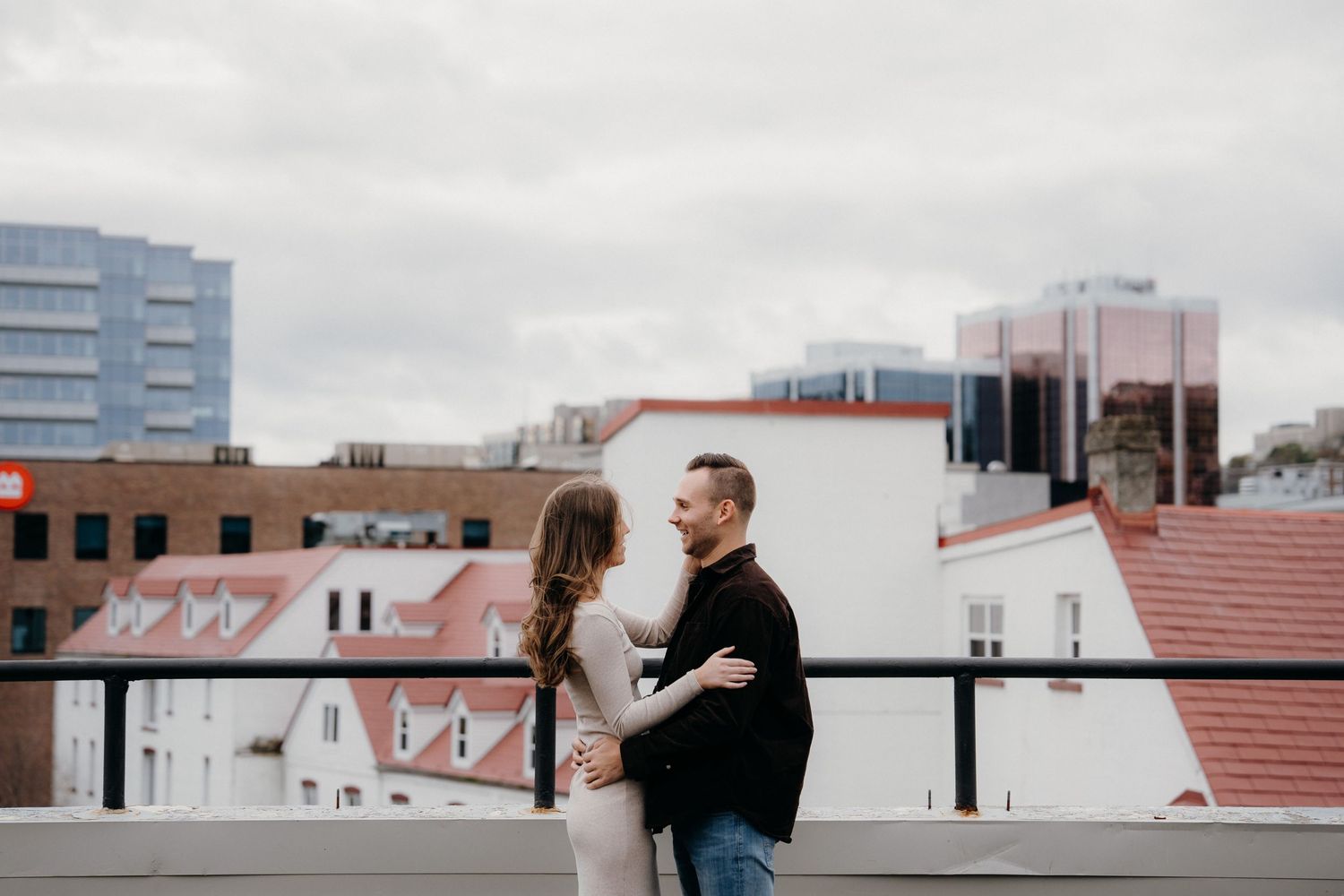 A couple embraces on an urban rooftop with city buildings in the background on an overcast day.