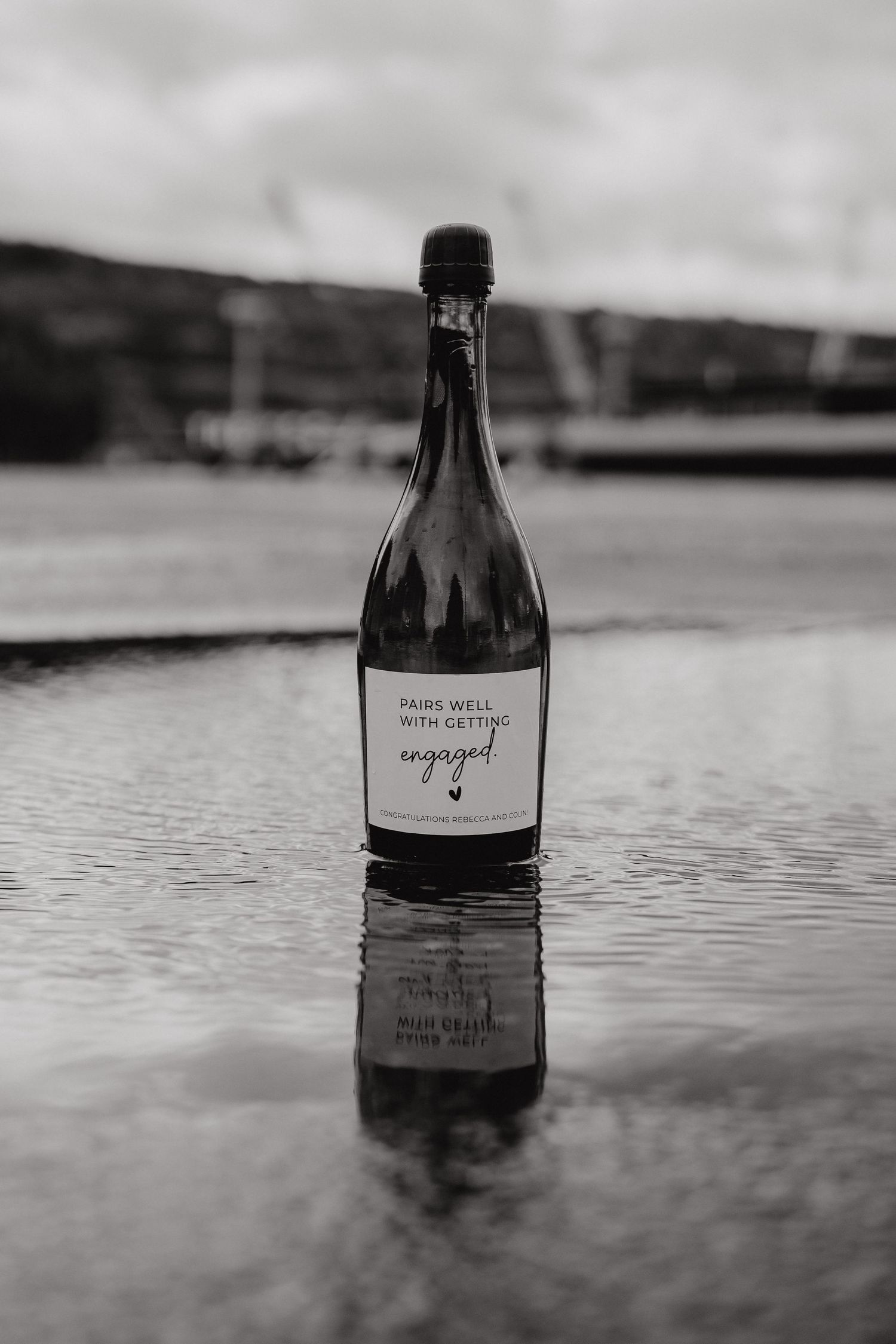 Black and white photo of wine bottle with label reflecting in shallow water.