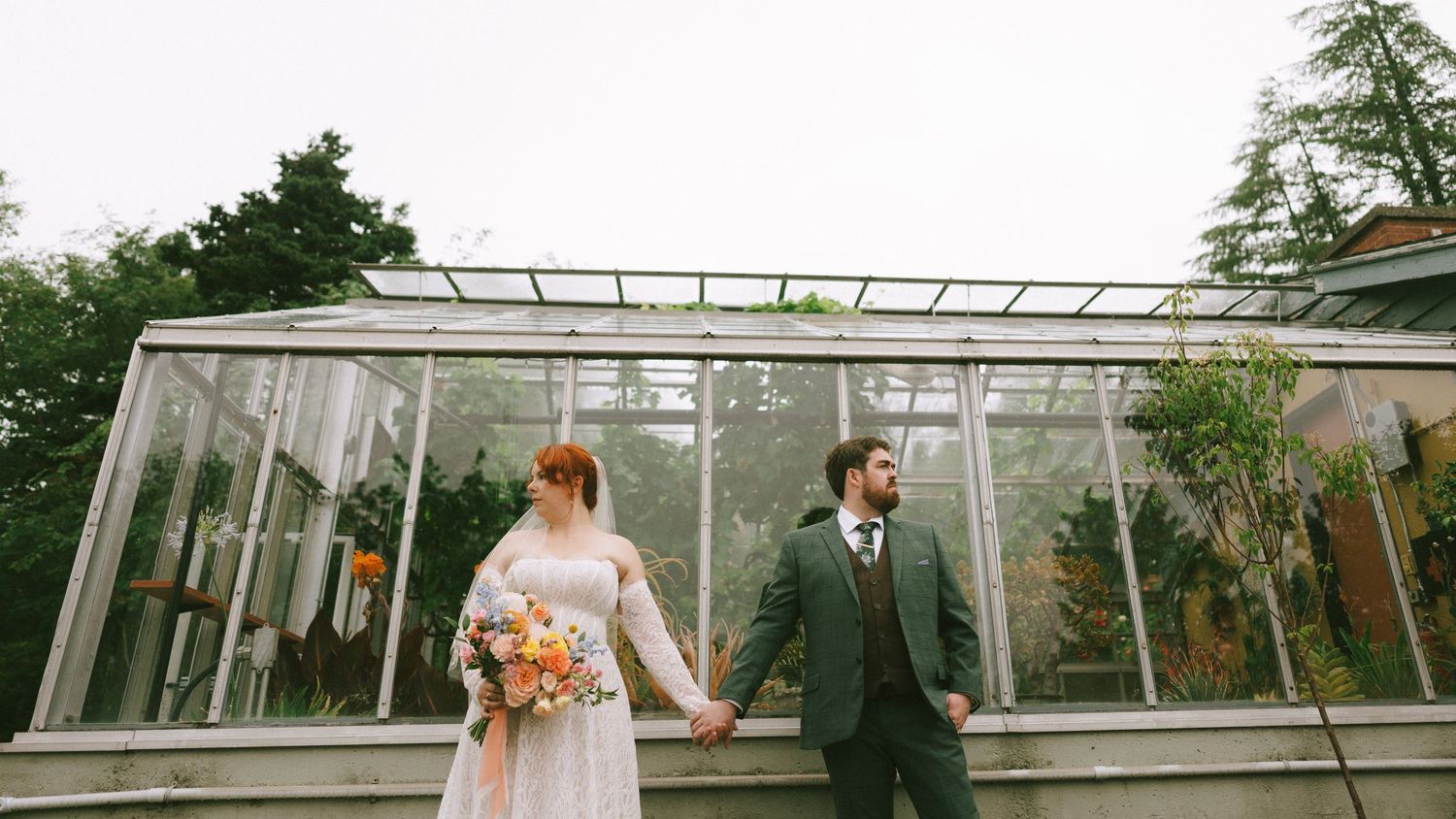 Bride and groom holding hands in front of a glass greenhouse on their wedding day.