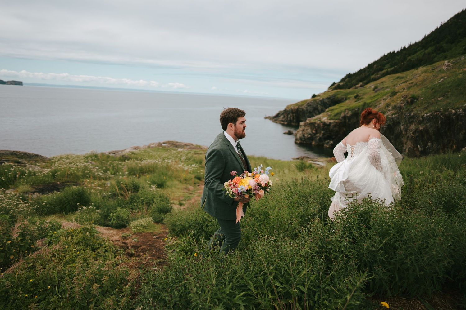Bride and groom walk through lush coastal greenery with ocean views on their wedding day.