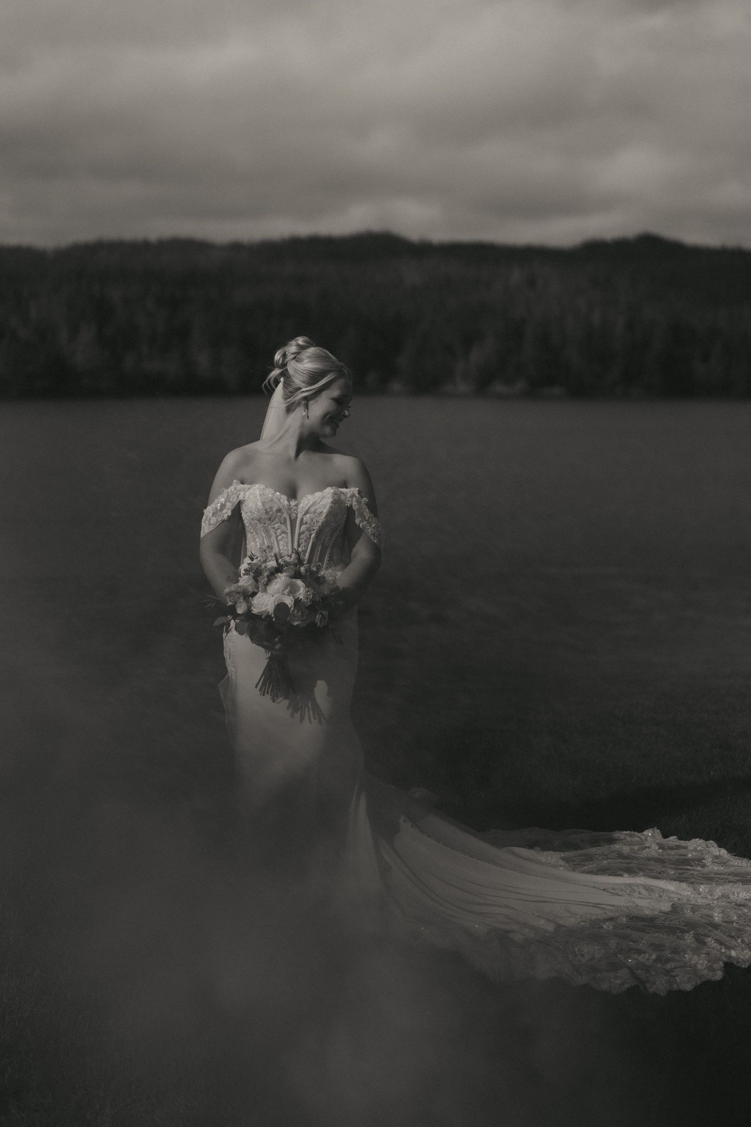 Black and white photo of a bride in an off-shoulder lace gown holding a bouquet, standing by a serene lake.