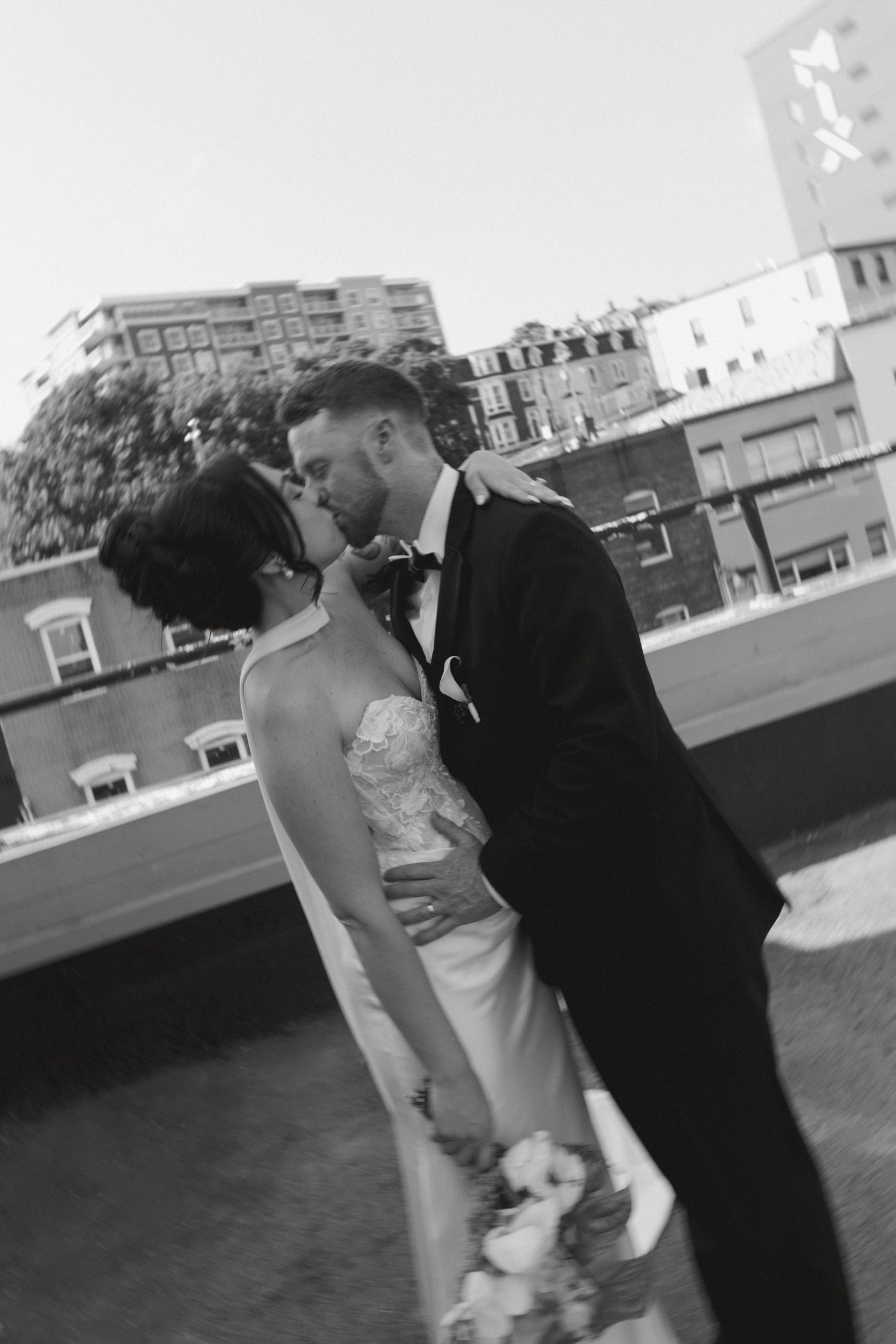 Black and white photo of a bride and groom sharing a kiss outdoors on their wedding day with city buildings in the background.
