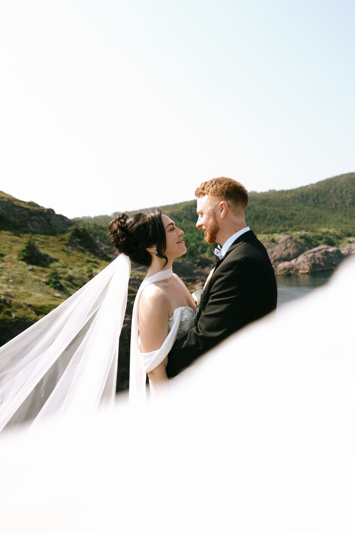 A bride and groom share a romantic moment outdoors, her veil flowing in the wind against a scenic mountain backdrop.