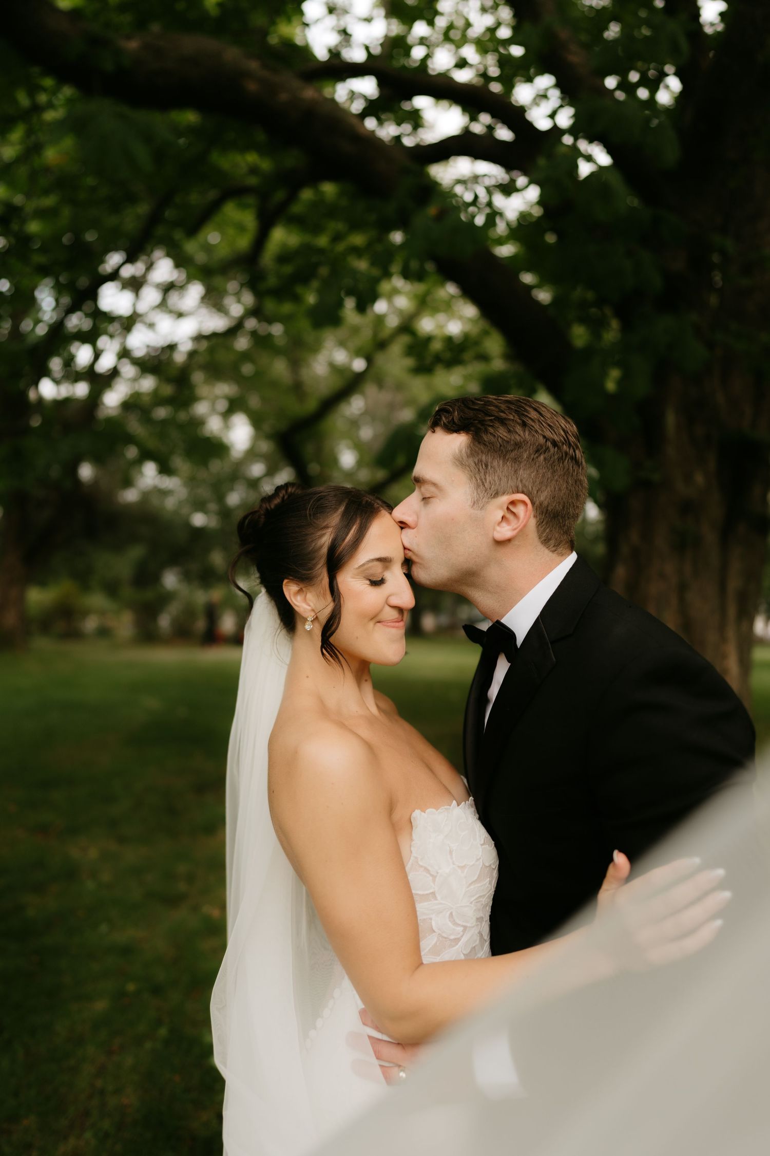 Groom in black tuxedo kissing bride in lace wedding dress and veil under lush green trees outdoors.