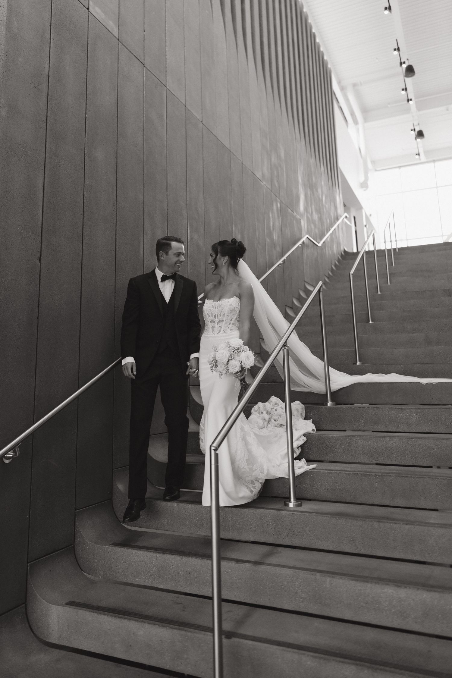 Black and white photo of a bride and groom sharing a romantic moment on grand stairs in an elegant venue.