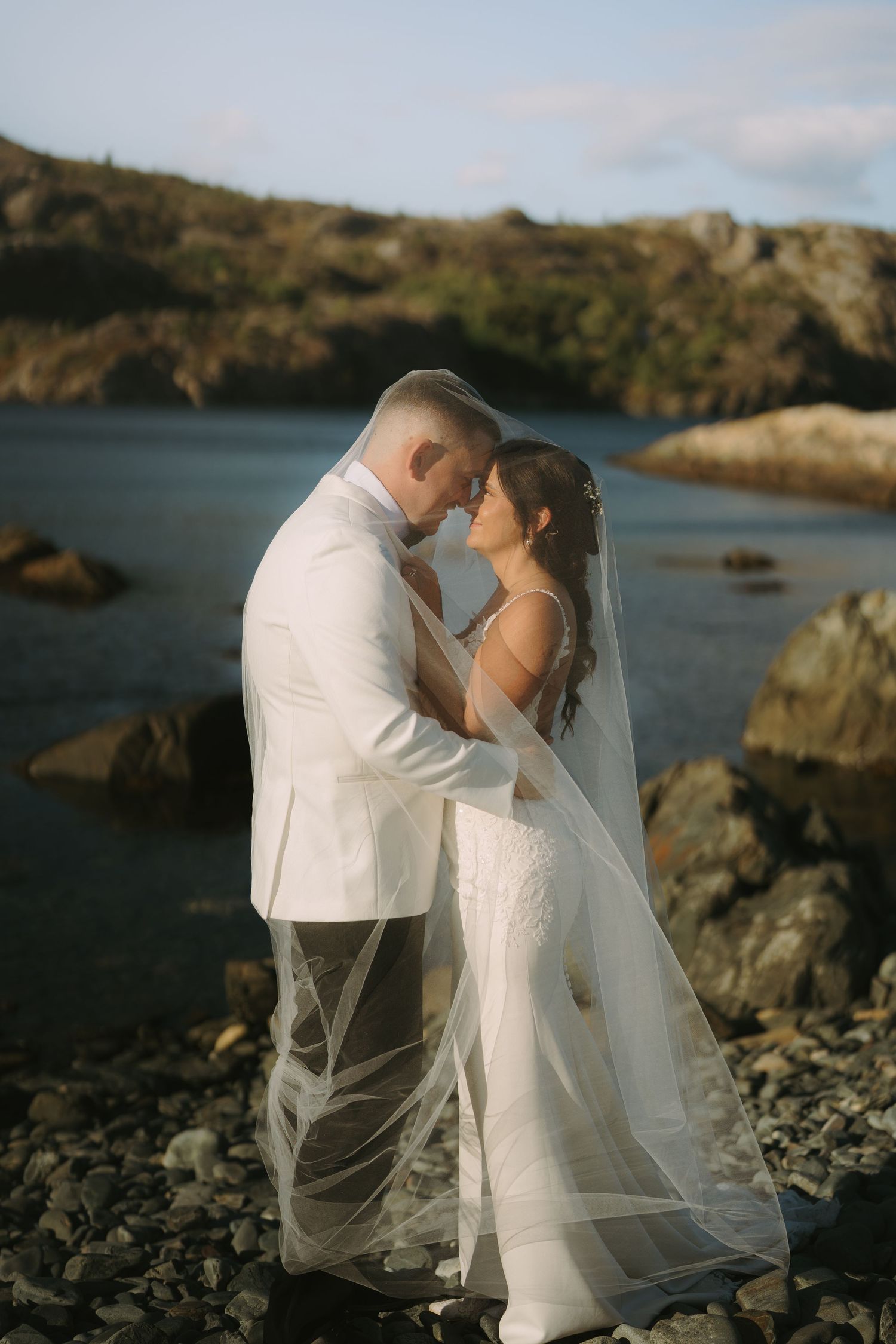 A couple shares an intimate moment in wedding attire on a rocky beach with cliffs and ocean in the background.