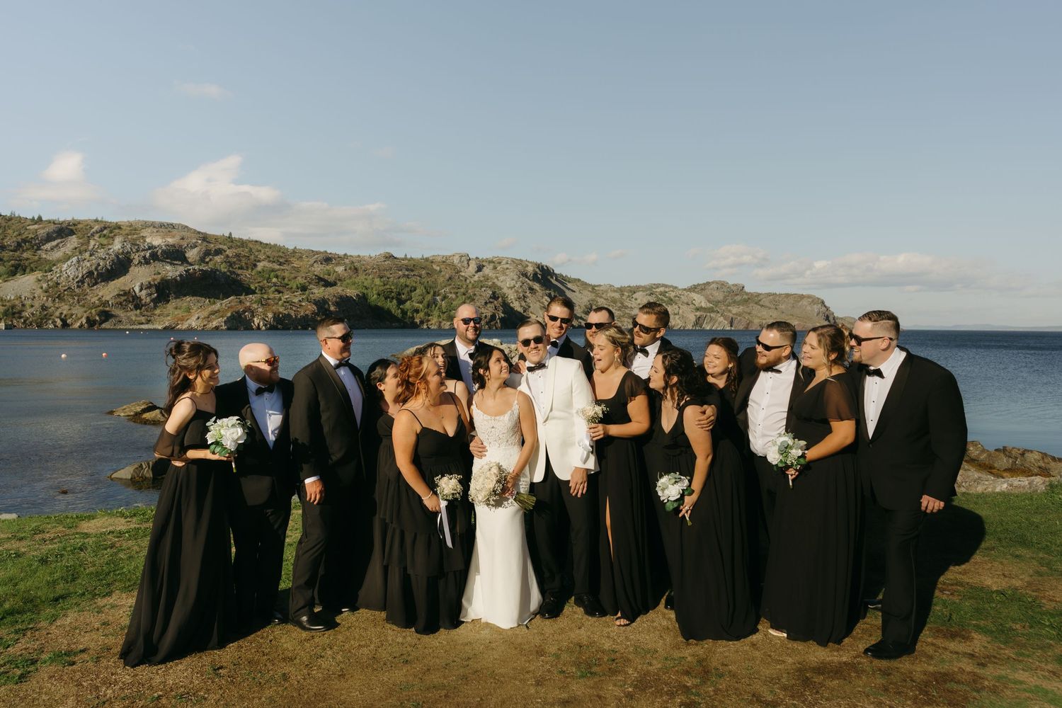 Wedding party in formal attire poses together against scenic coastal backdrop during outdoor celebration.