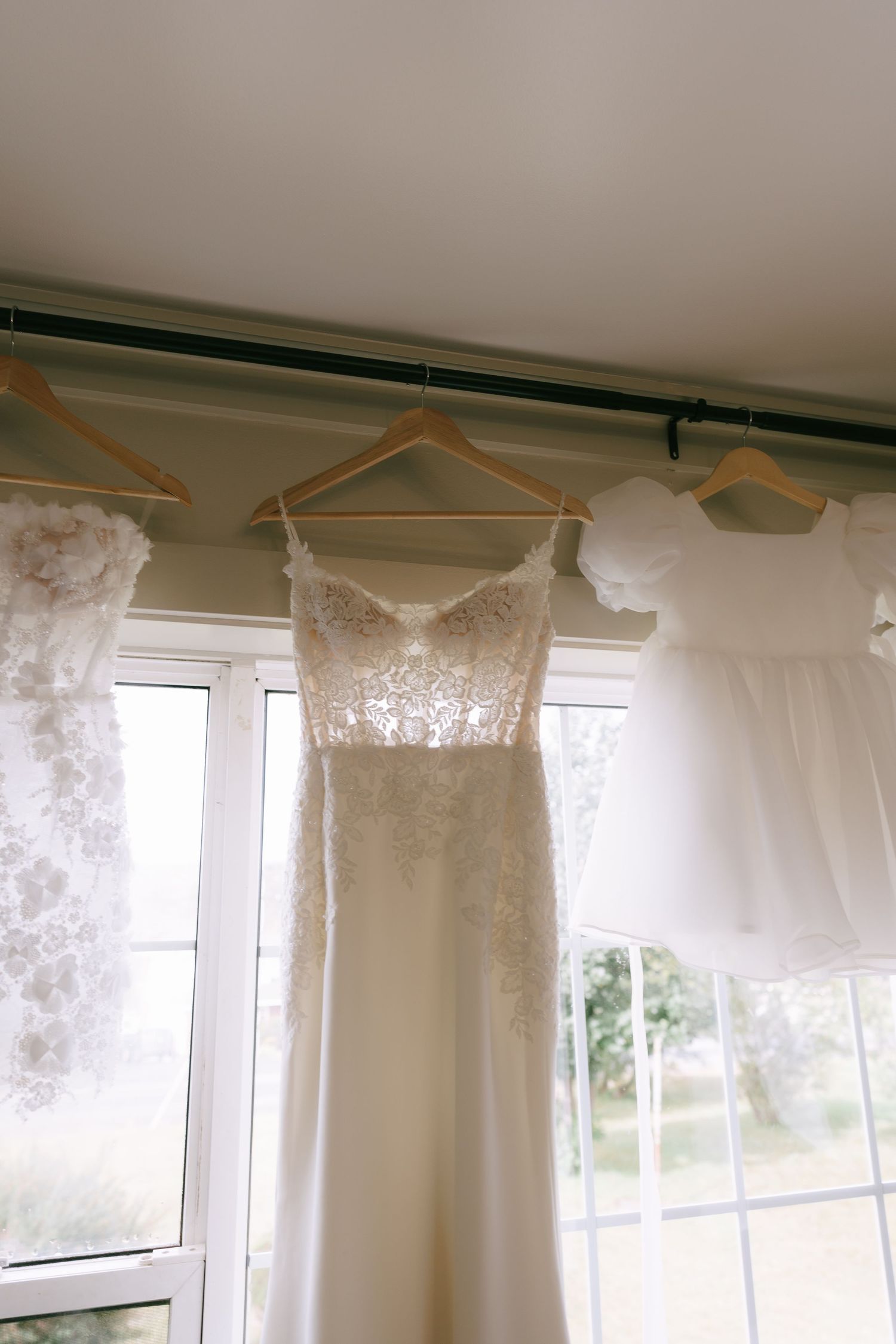 Several white lace wedding dresses hanging on hangers against a bright window in a bridal shop.