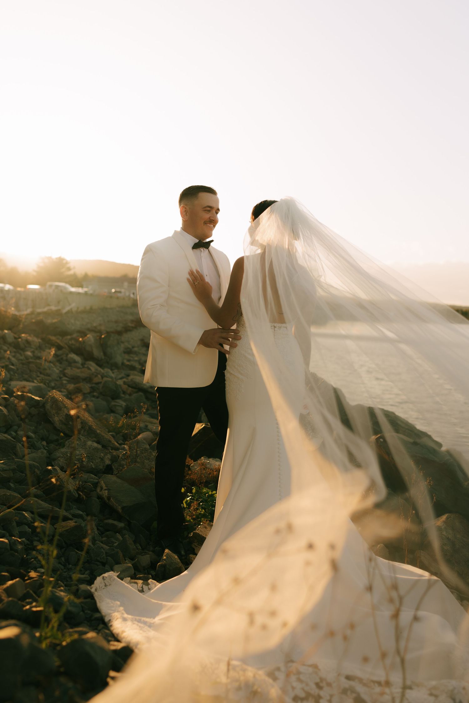 Romantic wedding couple poses on volcanic rocks during golden hour as bride's veil flows dramatically in the wind.