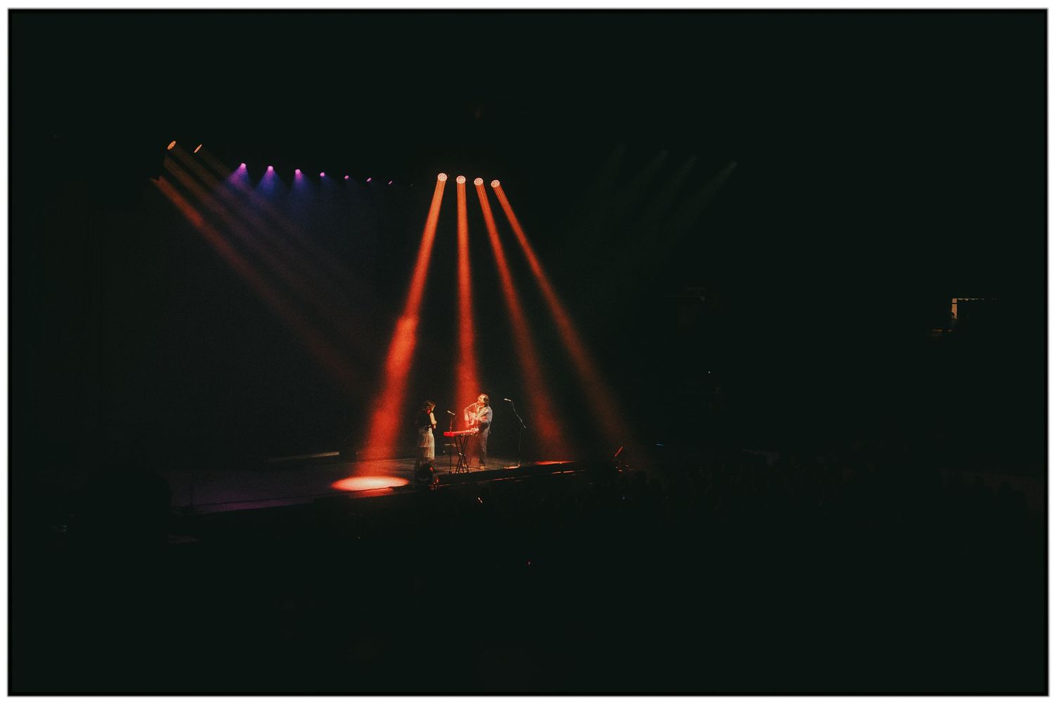 Stage performance illuminated by dramatic orange and purple spotlights creating beams of light in a dark concert venue.