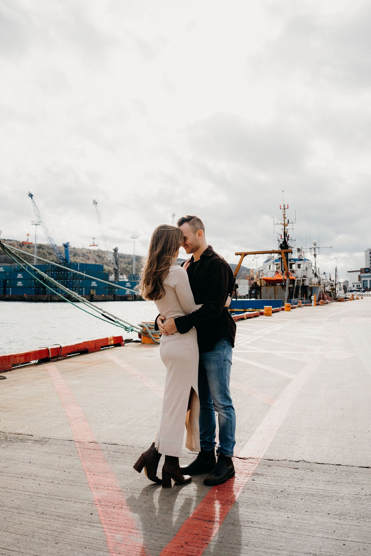 Elegant couple in romantic embrace on harbor dock with fishing boats and city skyline behind them.