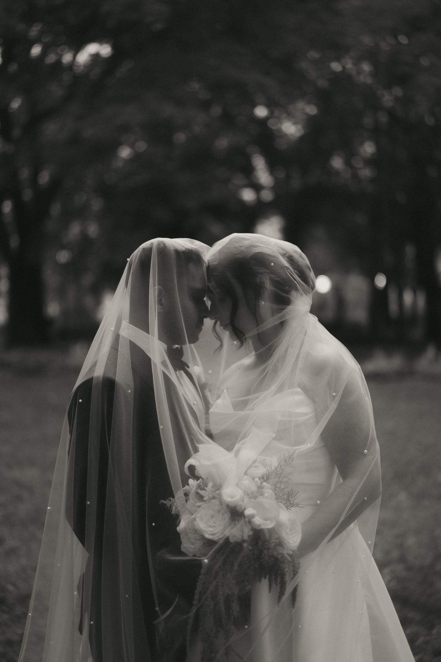 Black and white photo of two brides sharing an intimate moment under veils, one holding a bouquet.