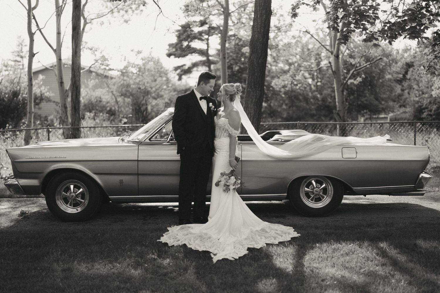 Black and white photo of bride and groom posing beside a classic convertible car outdoors.