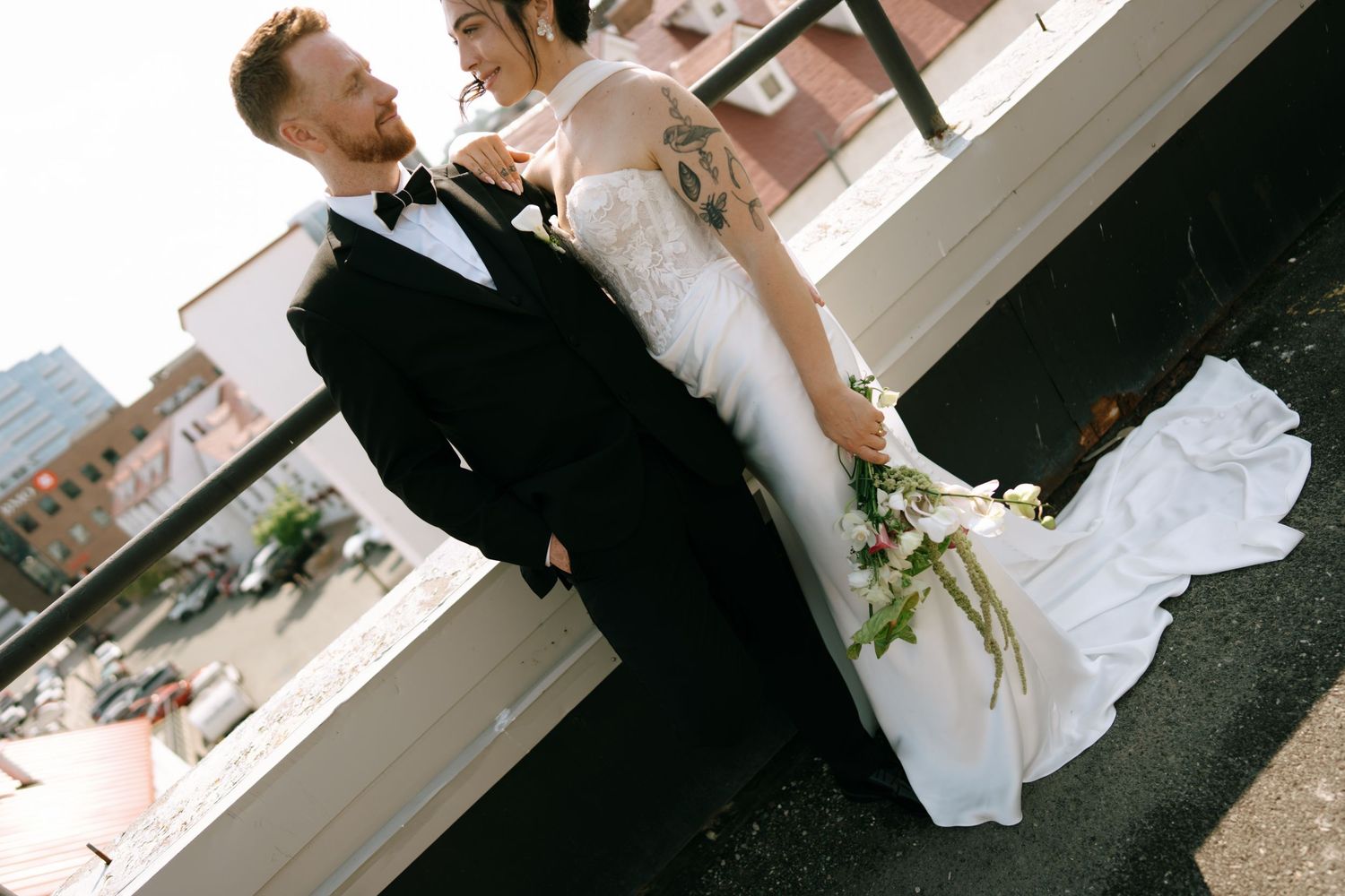 Bride and groom pose on rooftop, she wears lace gown holding bouquet, he wears black tuxedo, city skyline behind.