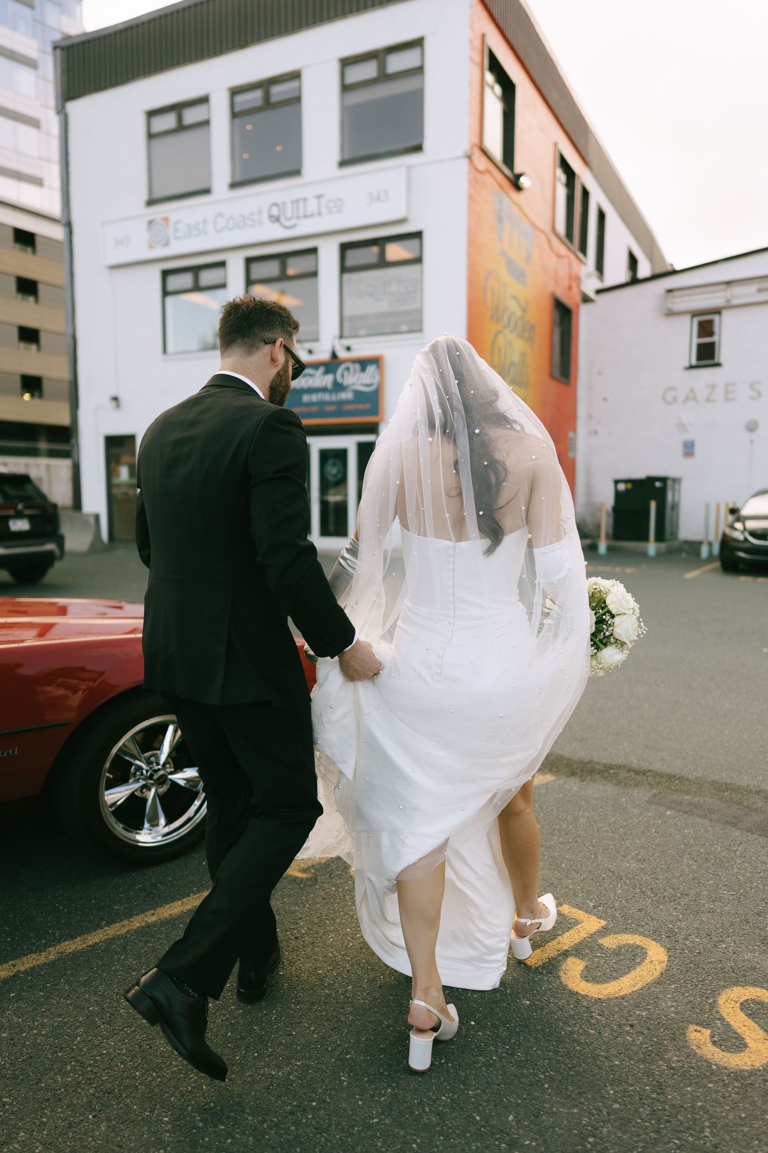 Bride and groom walking hand in hand through a parking lot, bride in white gown with veil holding bouquet.