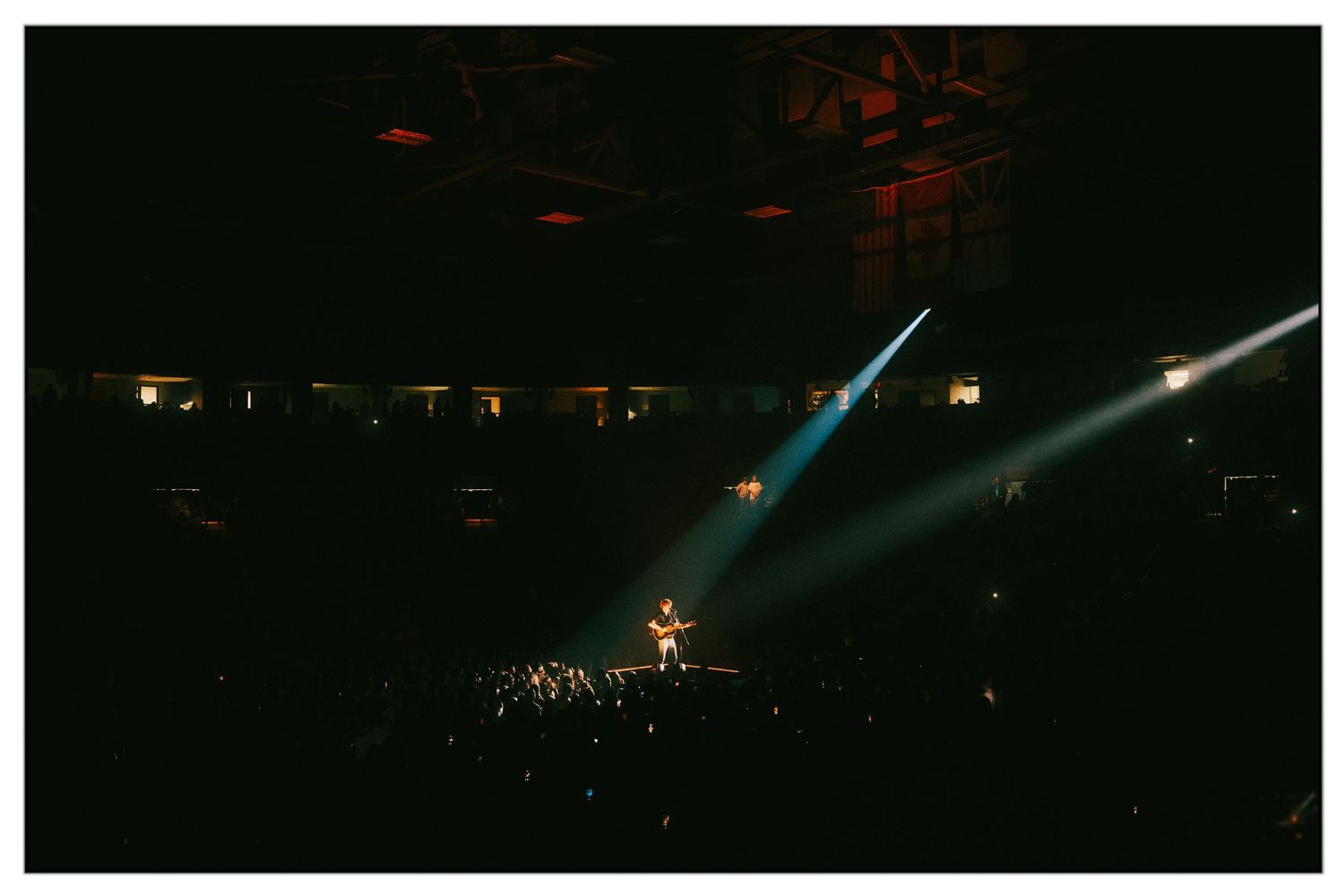 Concert venue illuminated by dramatic spotlights shining down on stage with crowd silhouetted in darkness.