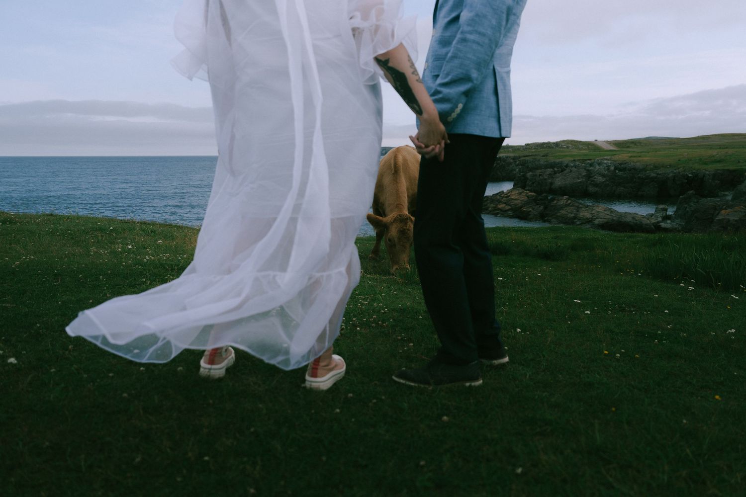 A couple in wedding attire walks along a scenic coastal cliff with flowing white dress in the breeze.