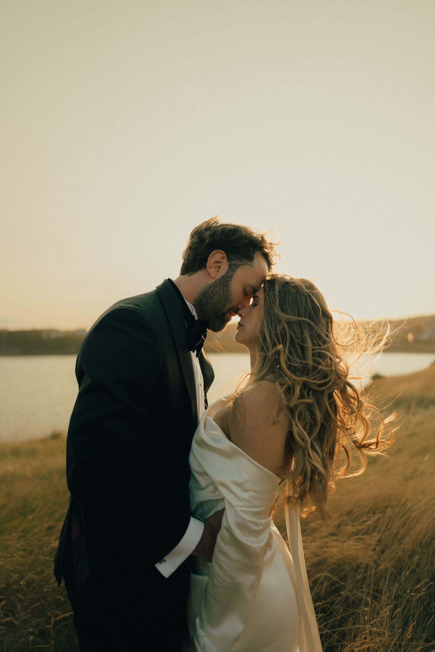 A romantic couple shares an intimate sunset moment in a golden field by the water.