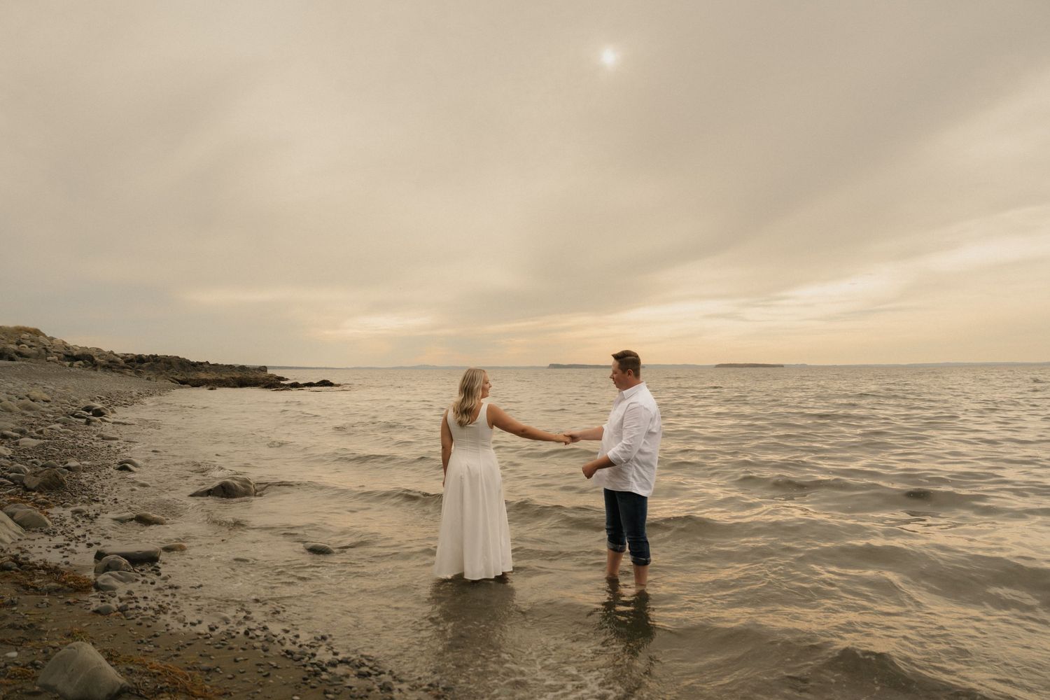 A couple embraces in shallow ocean water as the sun sets behind them creating a dreamy coastal scene.