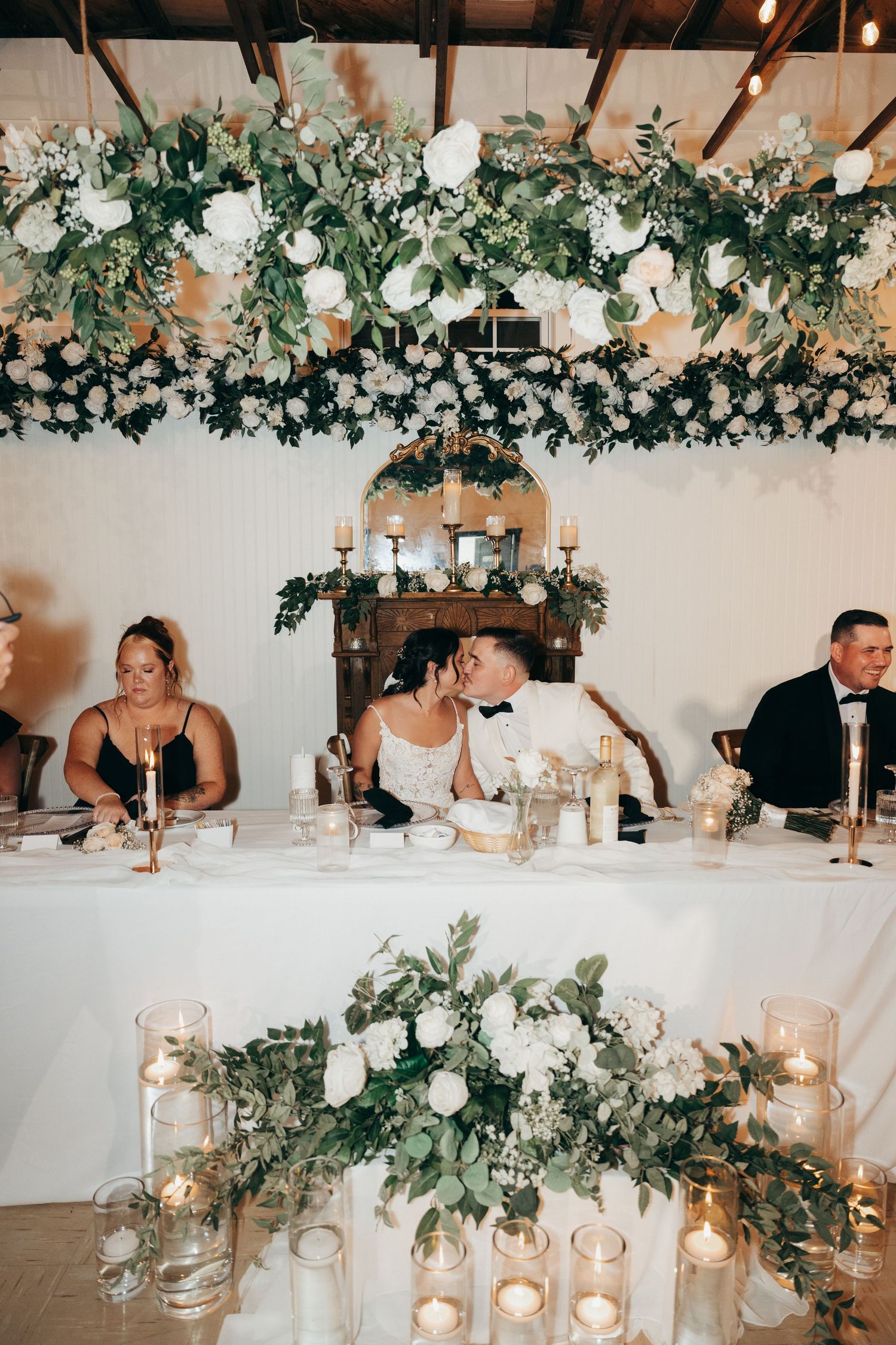 Wedding reception table decorated with elegant white floral arrangements, greenery and glowing candles in a bright venue.