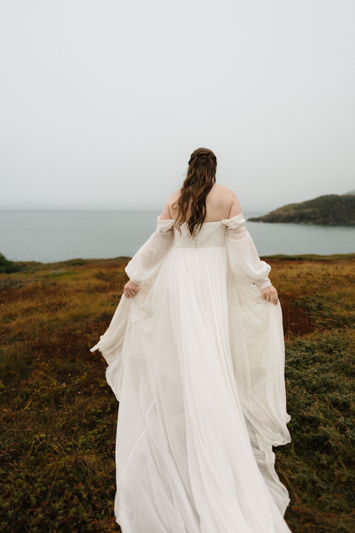 A flowing white wedding dress billows in the wind against a moody coastal landscape on a cloudy day.