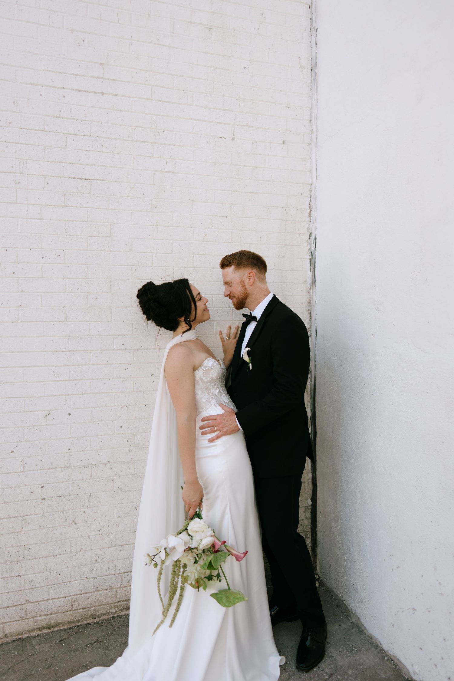 Bride in white satin gown holding bouquet faces groom in black tuxedo against white brick wall.