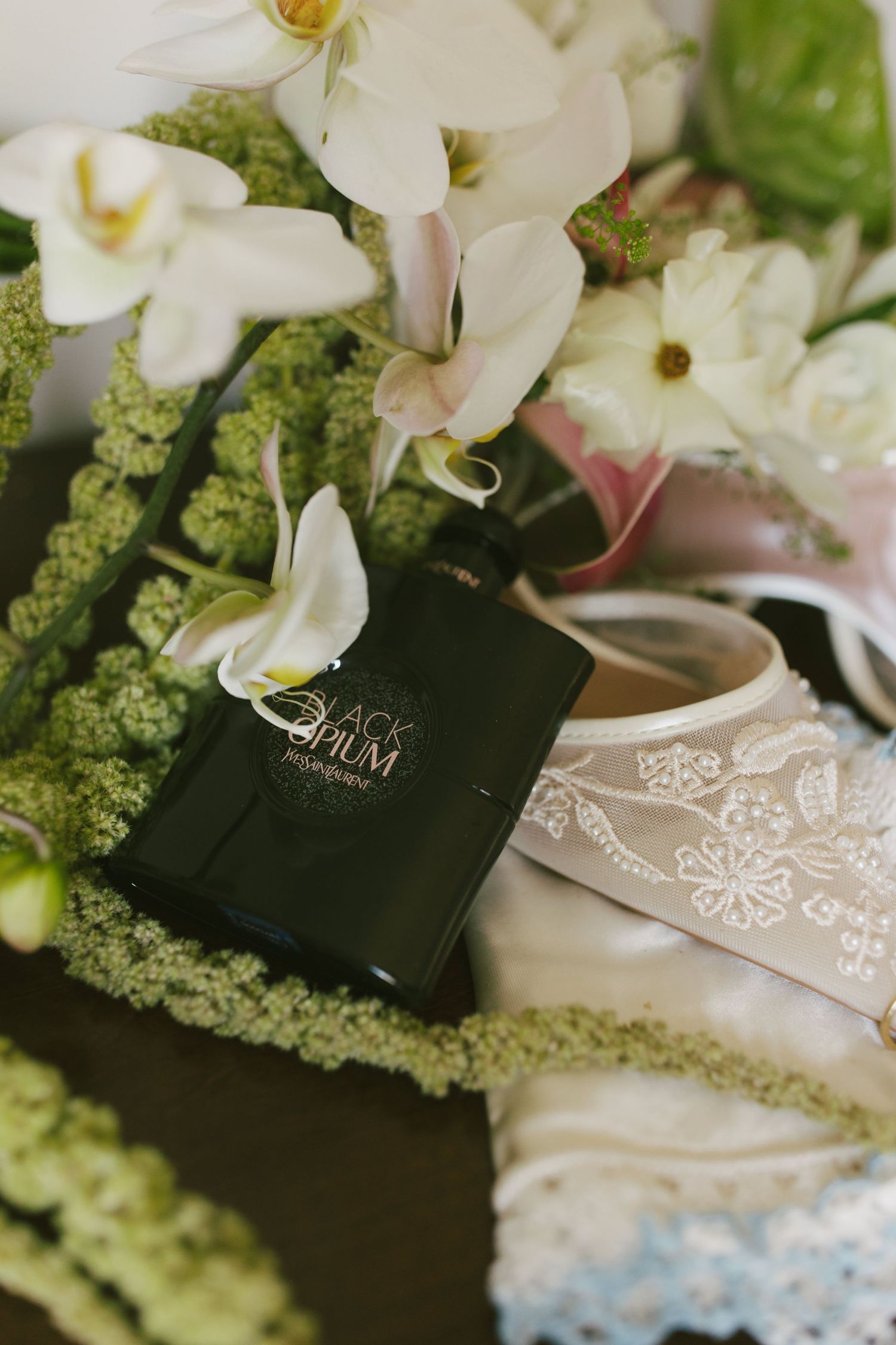 Black Opium YSL perfume bottle nestled among white orchids, green florals, and delicate lace bridal accessories.