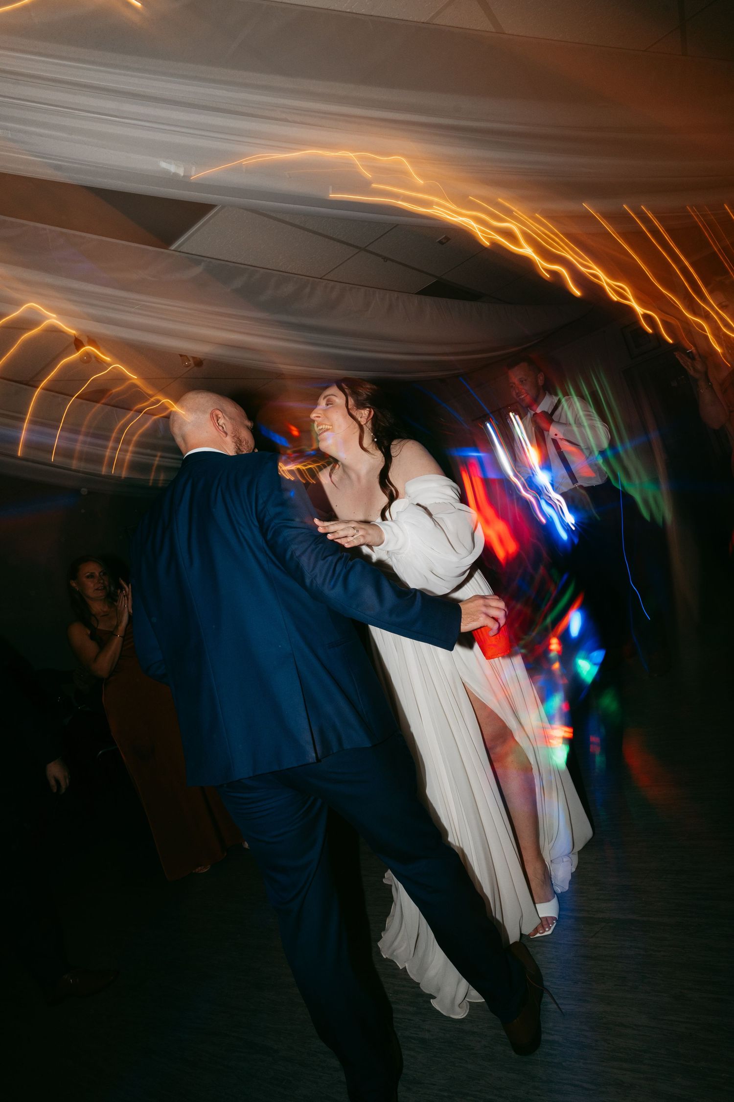 Couple dancing energetically at a wedding reception with colorful light trails and motion blur in dark venue.