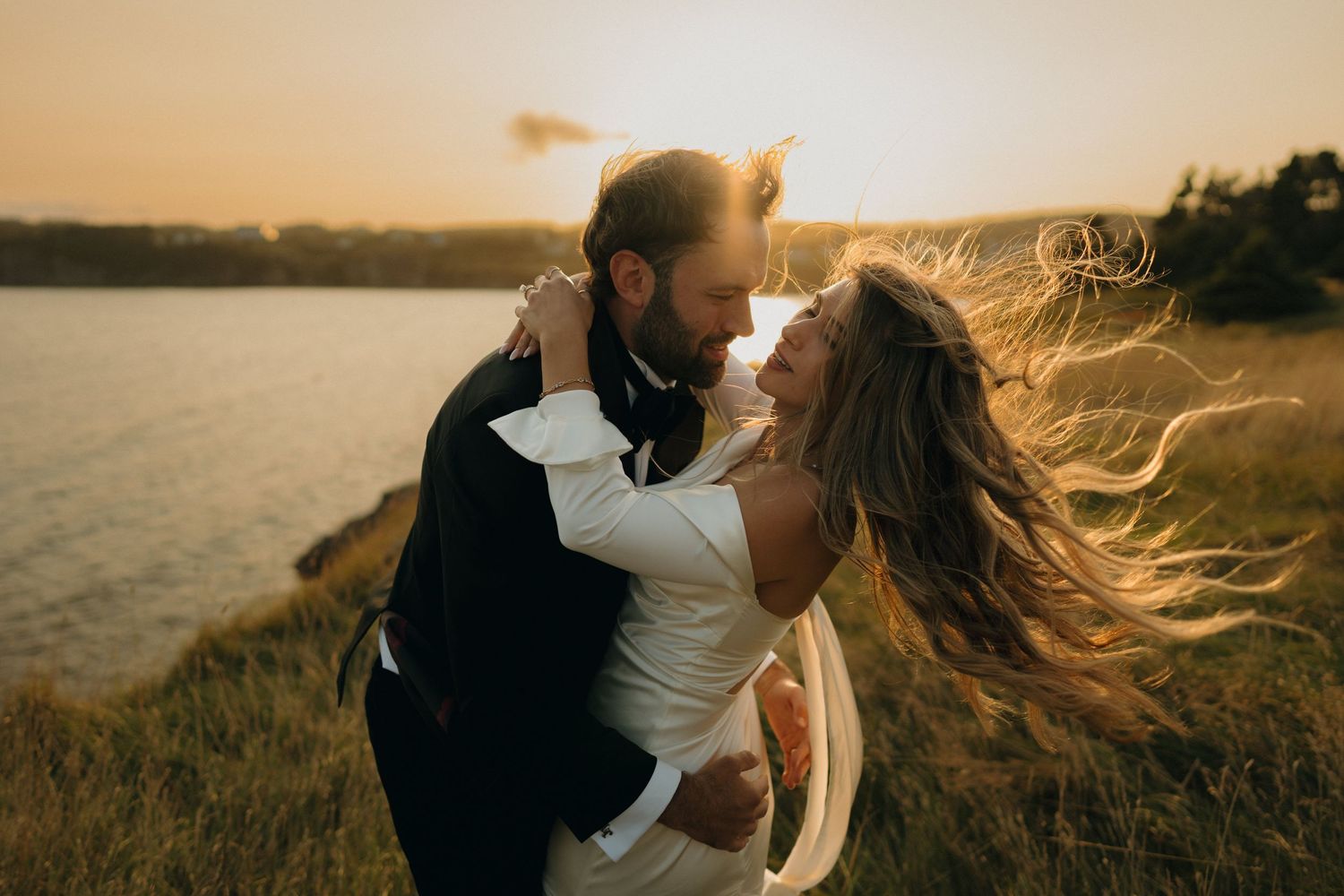 A romantic couple embraces at sunset by the water as wind blows through their hair in a series of dreamy photos.