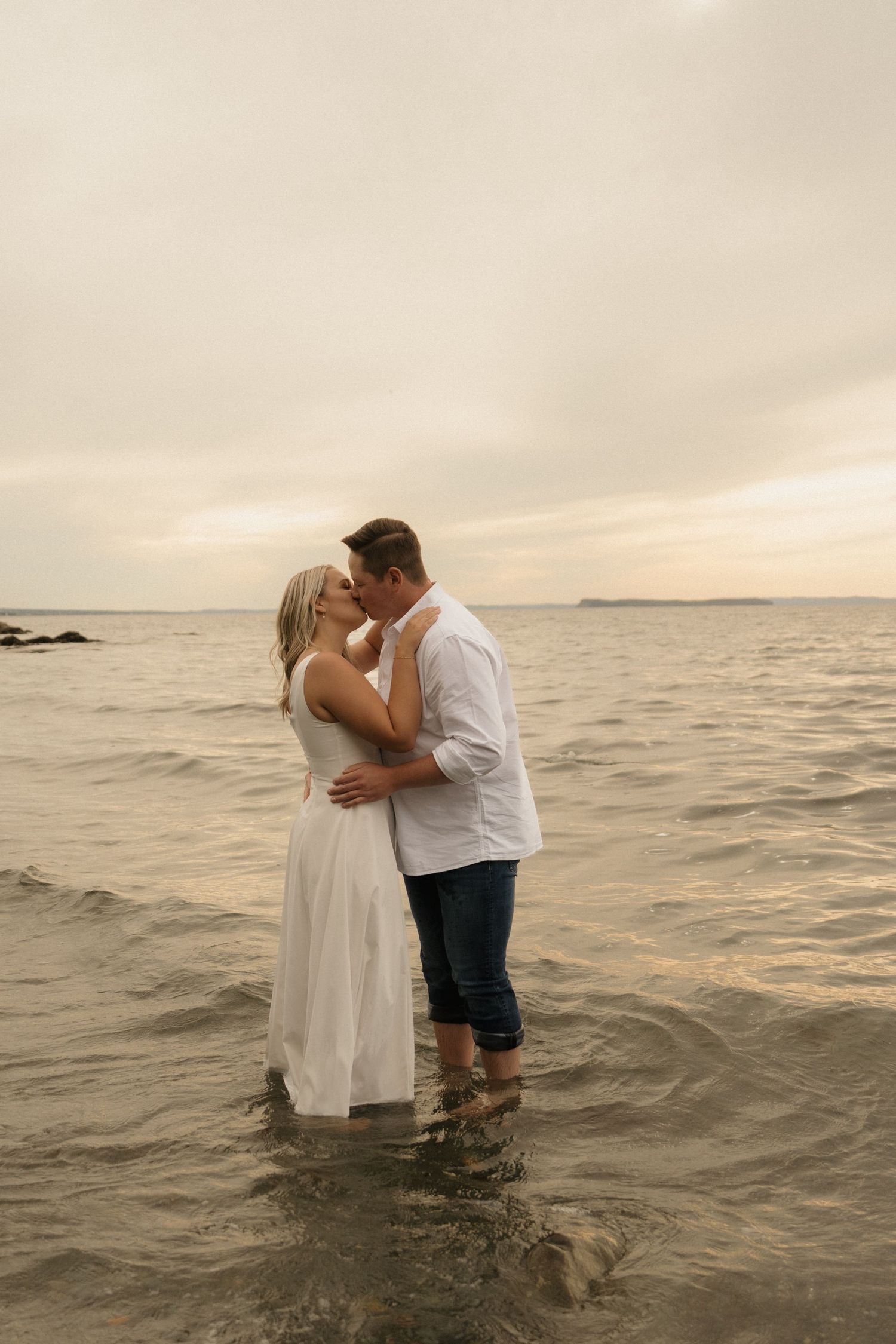 A couple embraces while standing in shallow ocean water during sunset, creating a romantic coastal scene.