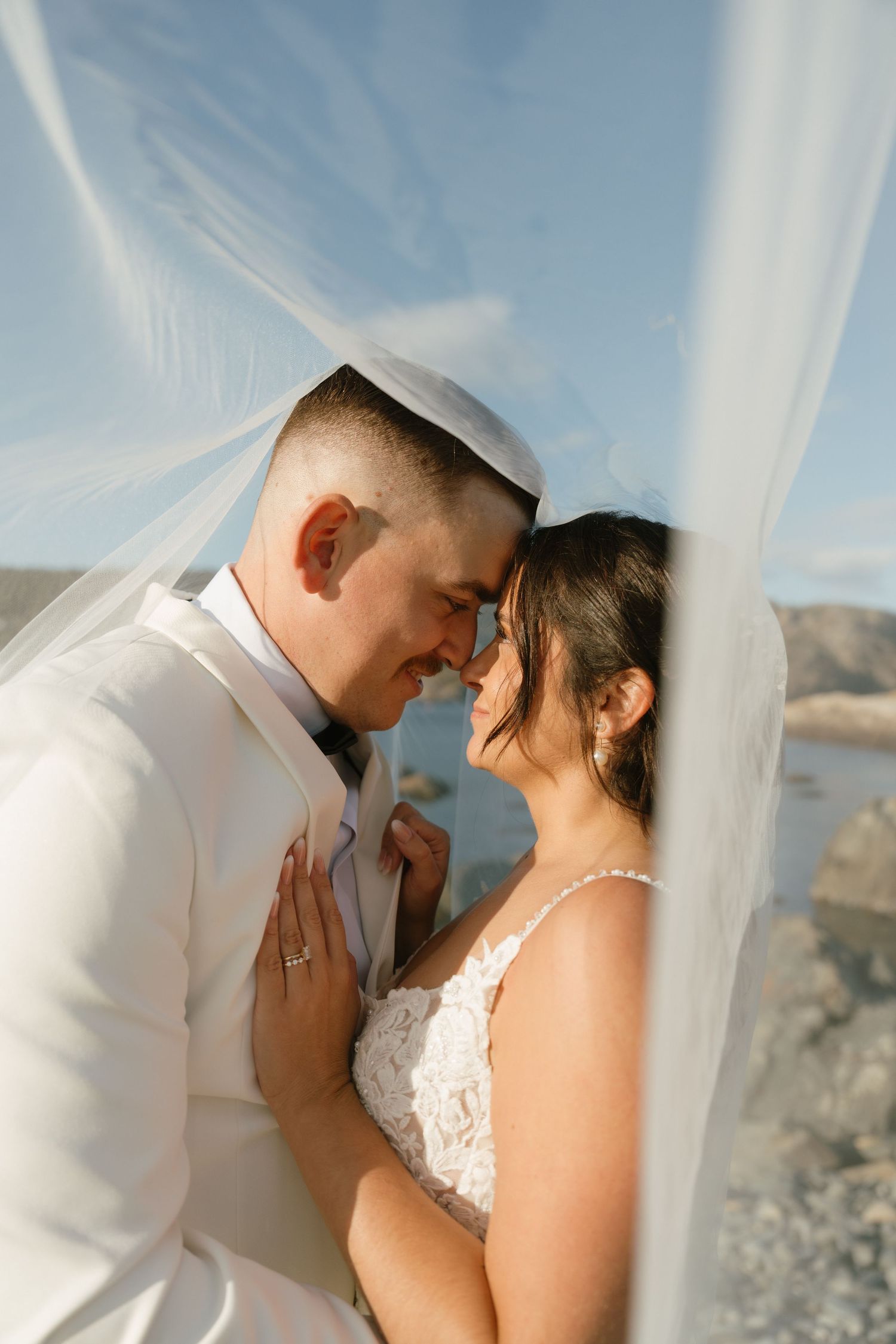Romantic wedding couple embraces behind flowing white curtains against a scenic mountain backdrop at sunset.