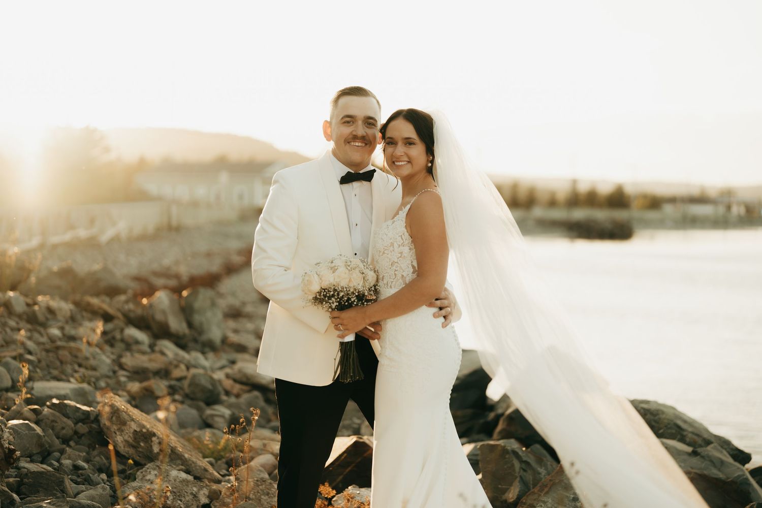 A couple in wedding attire embraces on a rocky beach during sunset, creating a romantic silhouette against the golden sky.