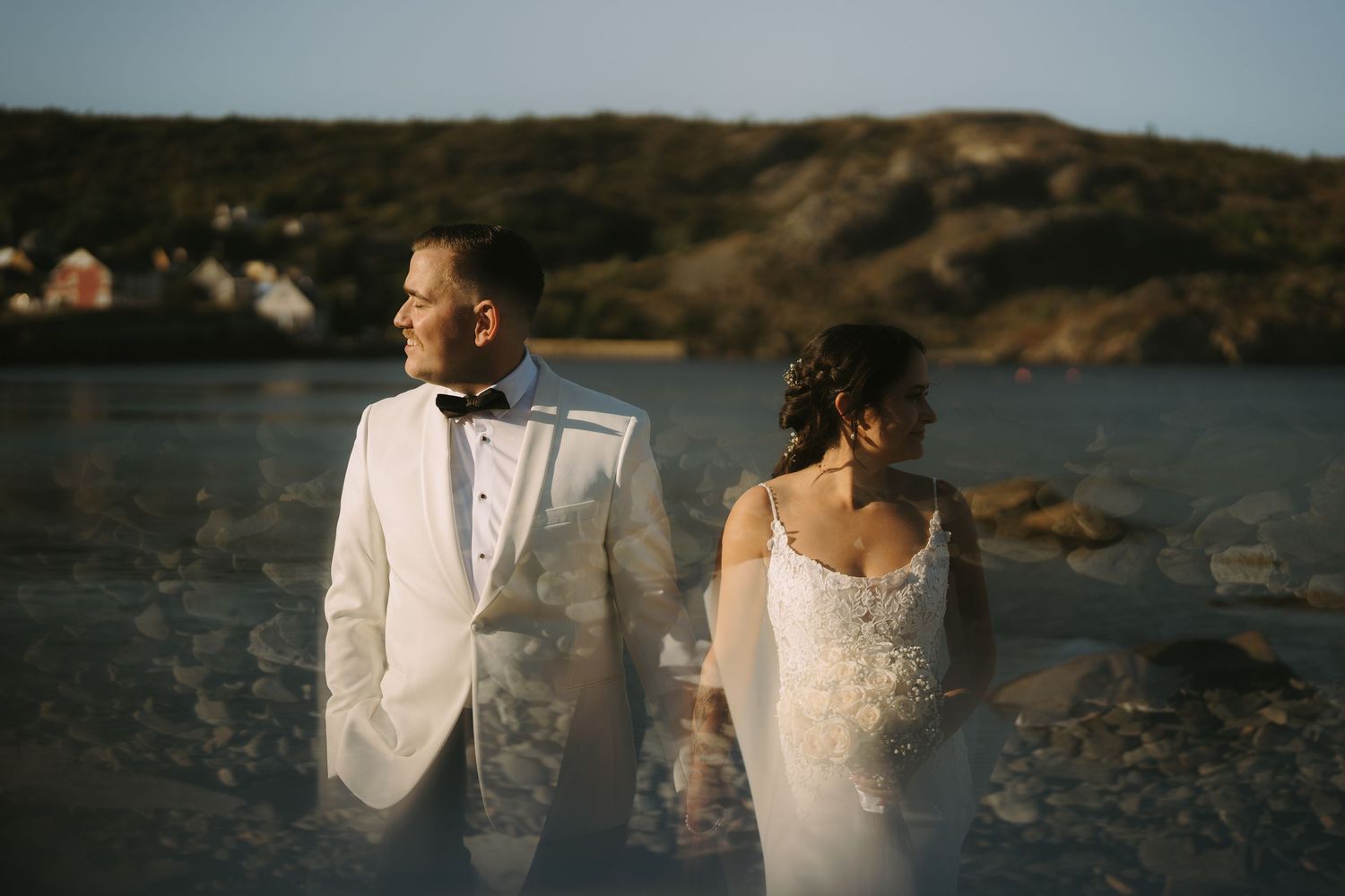 Couple in wedding attire embraces on rocky coastal shoreline during sunset photoshoot.